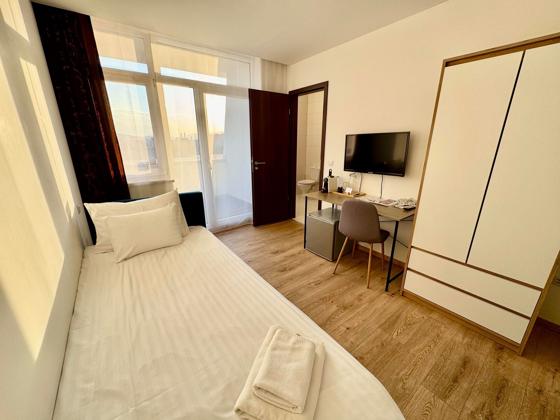A modern hotel room with a single bed, desk, TV, and large wardrobe. Natural light streams in.