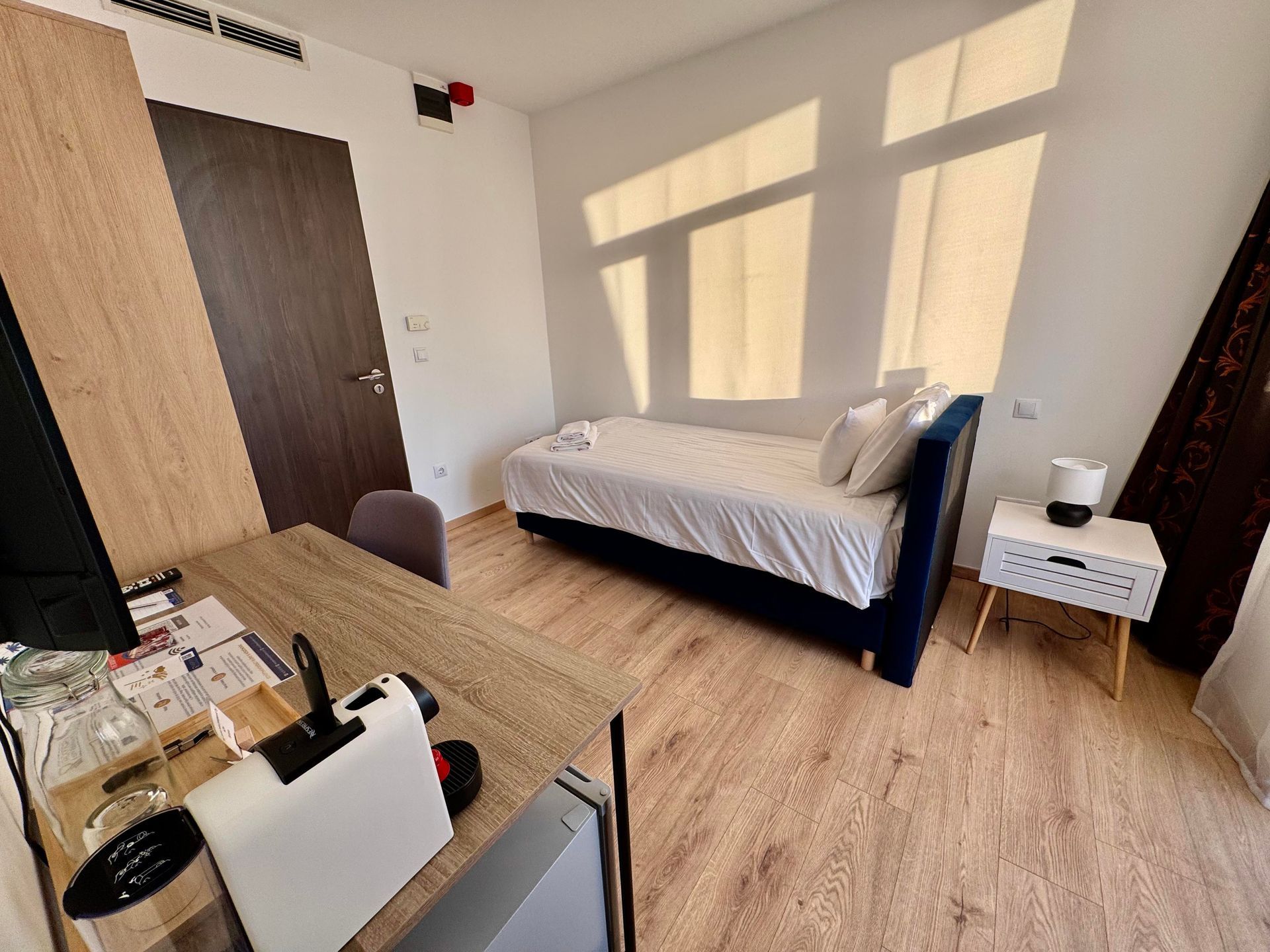 Bedroom with a single bed, desk with coffee machine, and a small nightstand. Light streams in.
