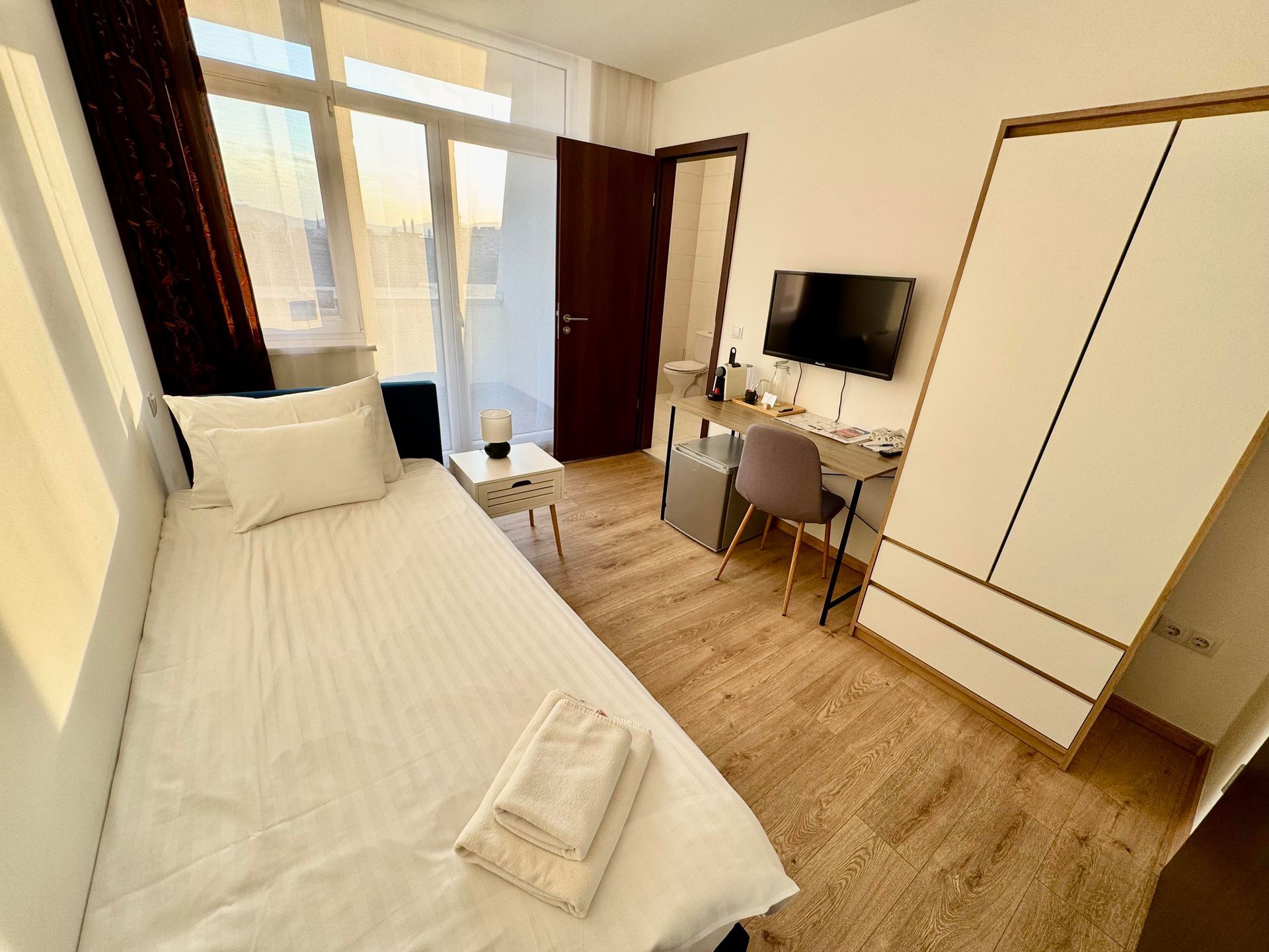 Small bedroom with a single bed, desk, and wardrobe. Sunlight streams in from a window.