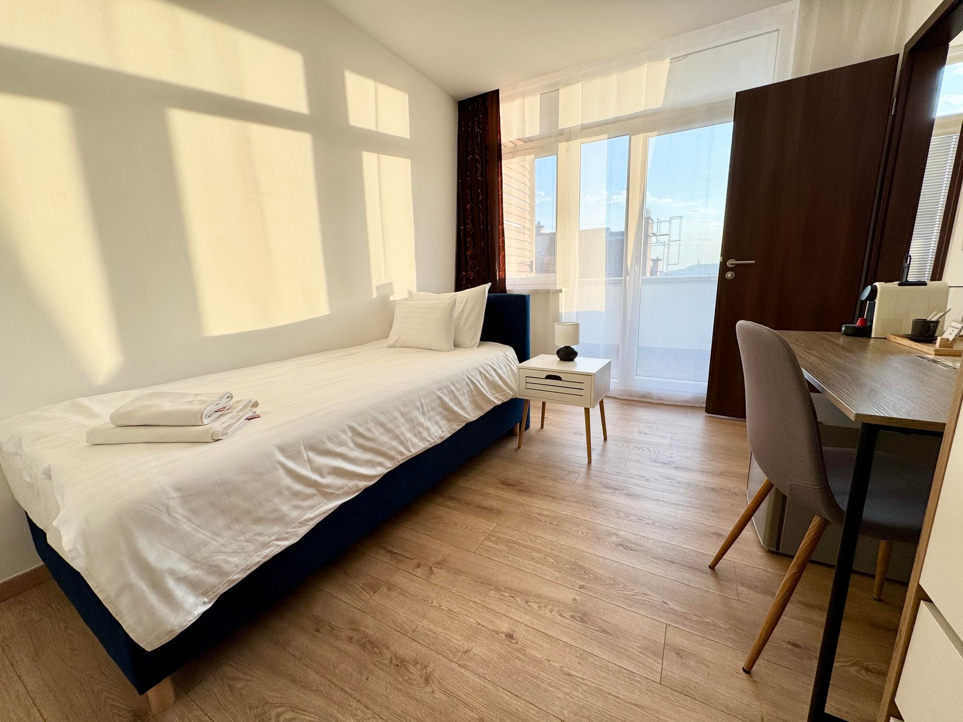 Bedroom with a single bed, desk, and balcony access. Sunlight streams through the window.