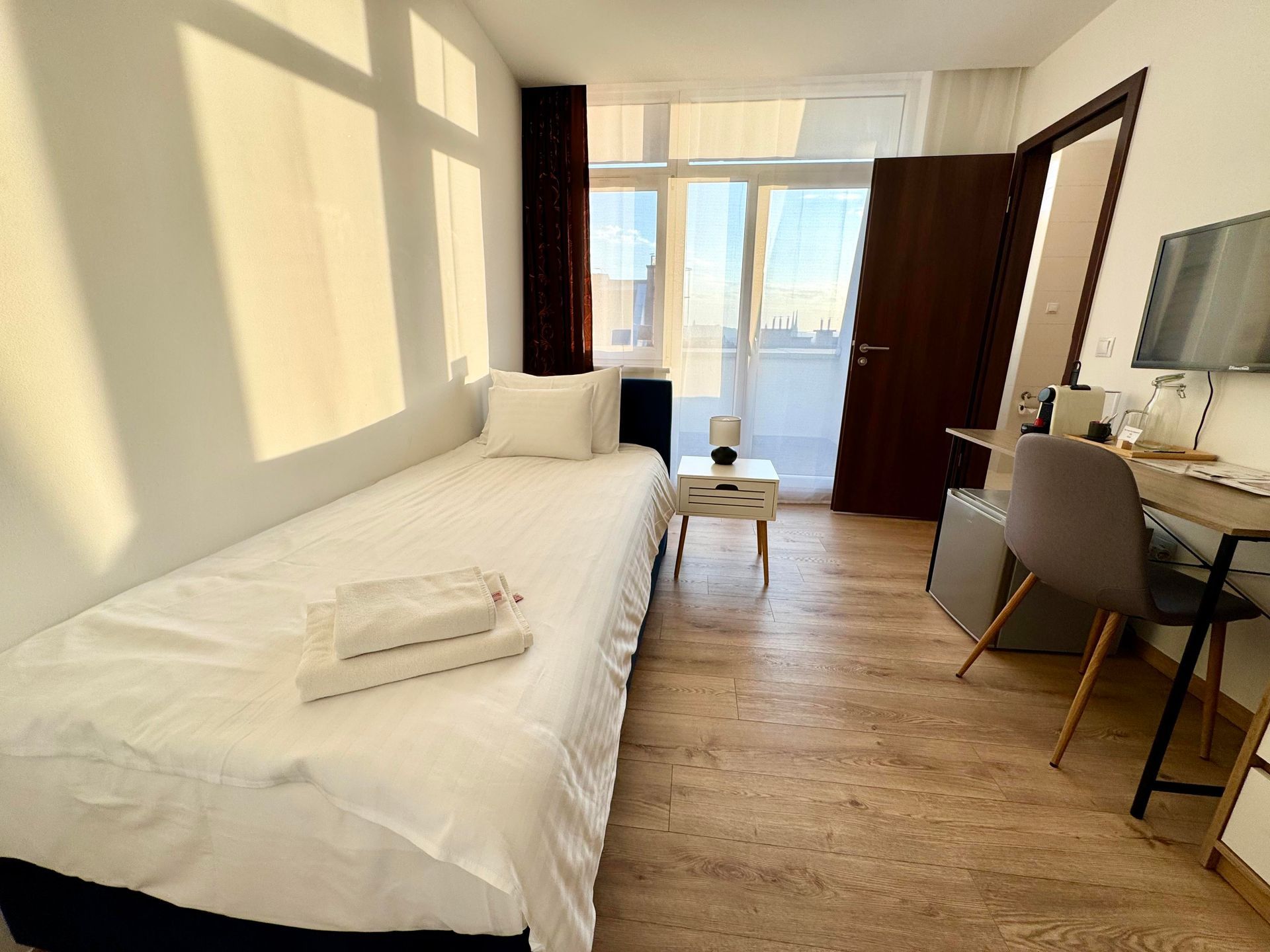 Bright hotel room with single bed, desk, and balcony door. Sunlight streams in.
