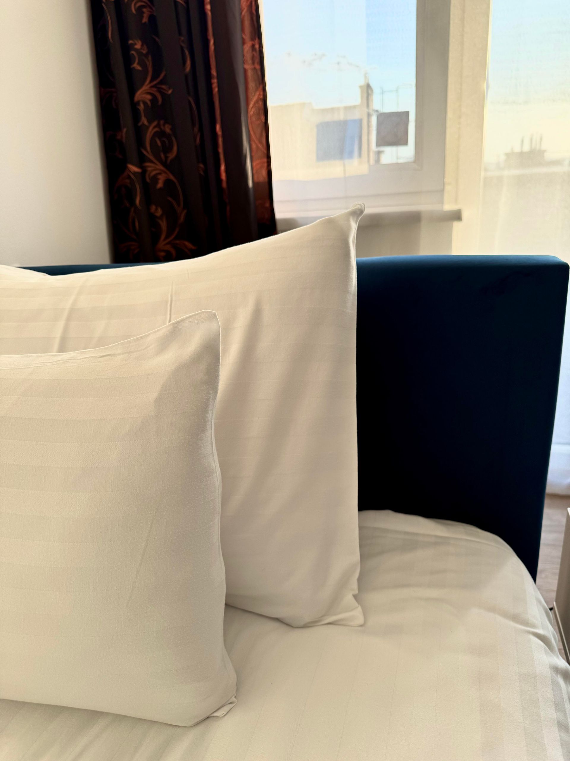 White pillows on a blue bed. Brown curtain and window in the background.