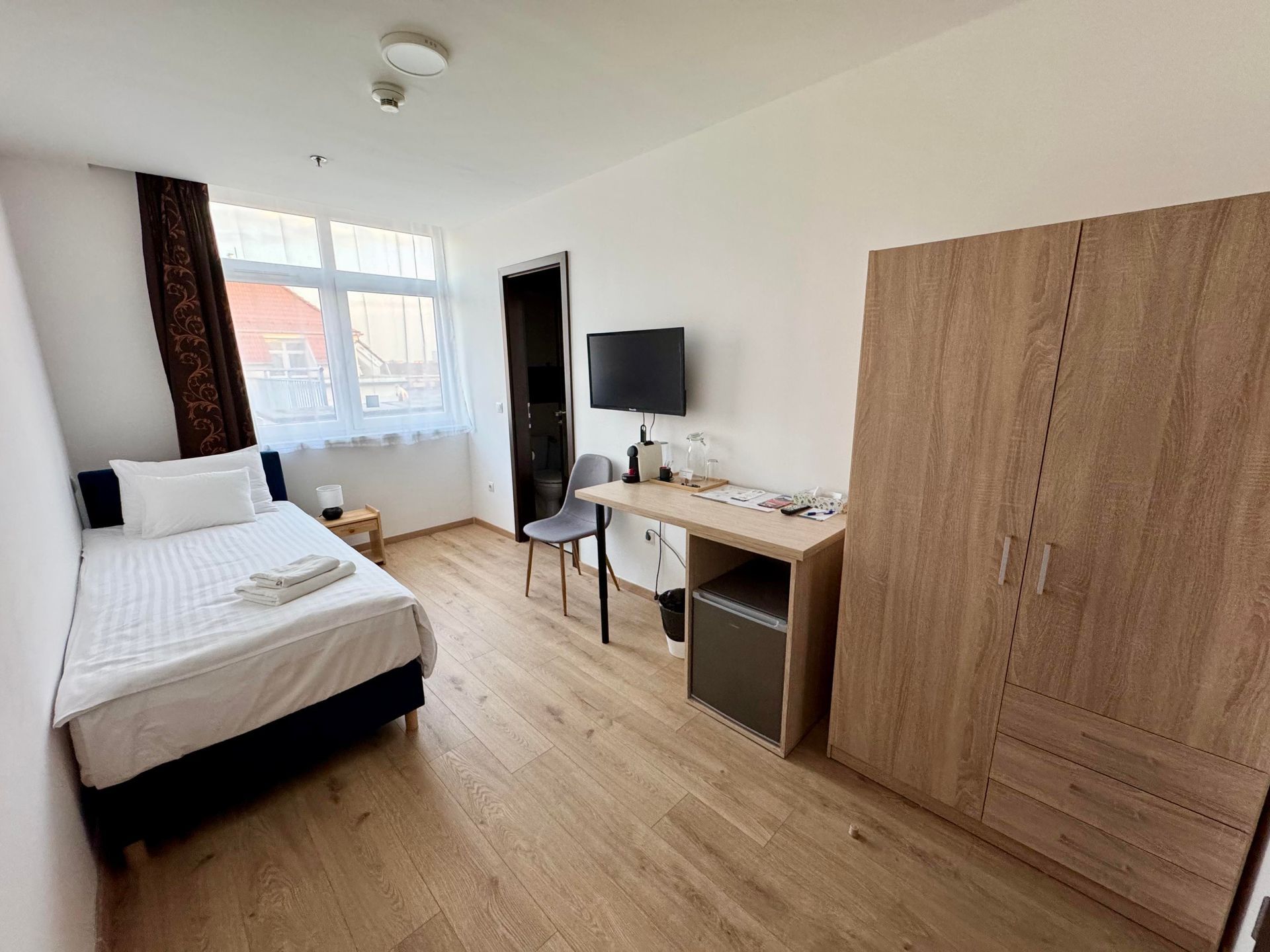 Small hotel room with bed, desk, wardrobe, TV, and window.