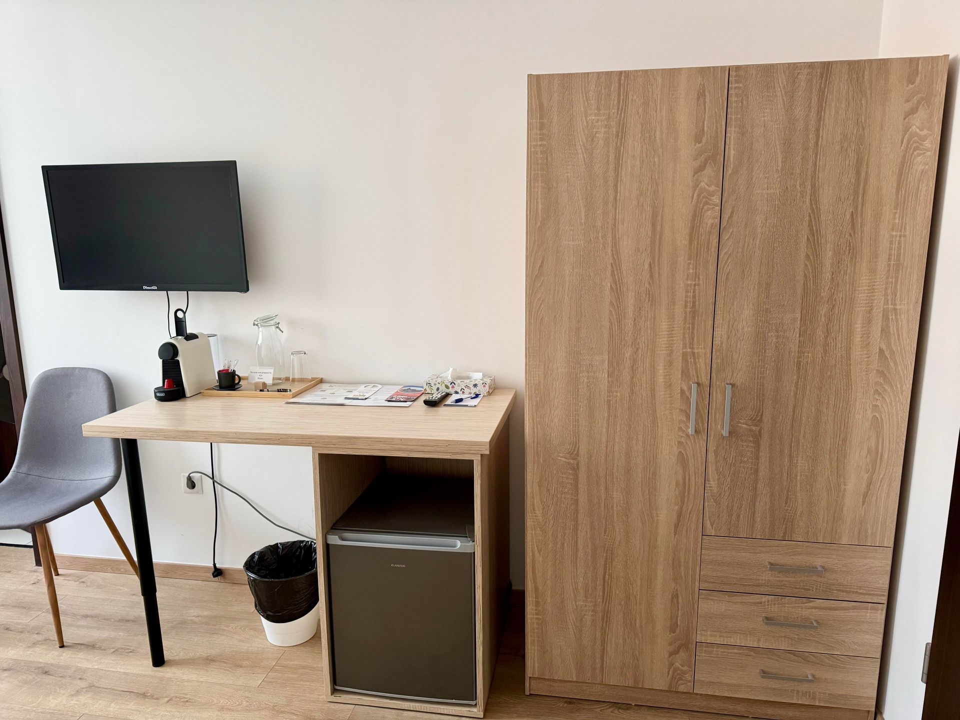 A room with a TV, desk, closet, and chair. A mini-fridge sits under the desk.