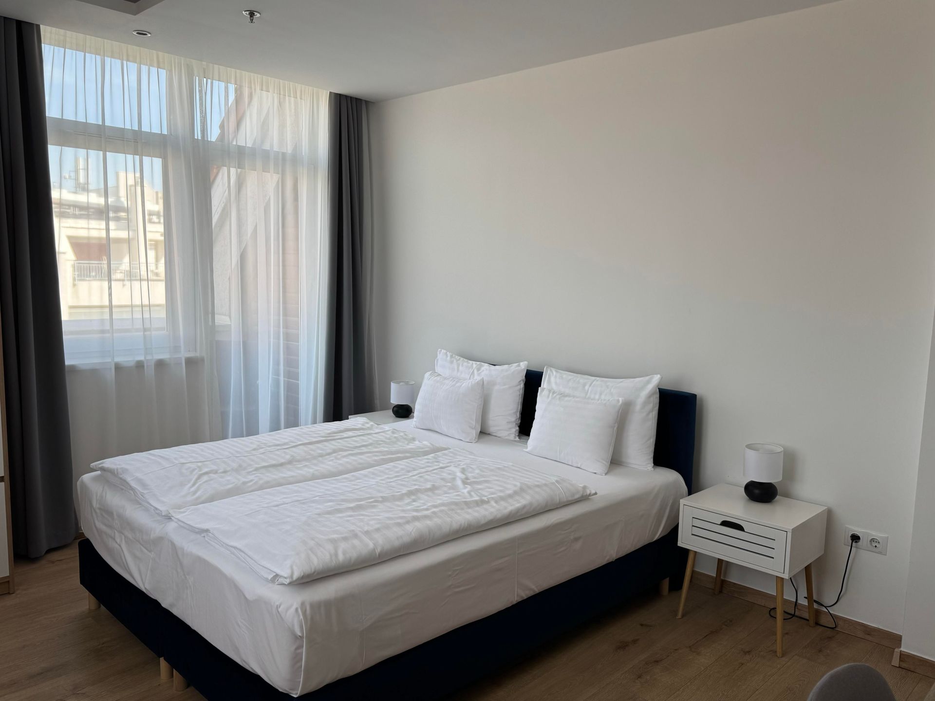 Bedroom with a bed, nightstand, window, and white walls. Dark blue bed frame, white bedding.