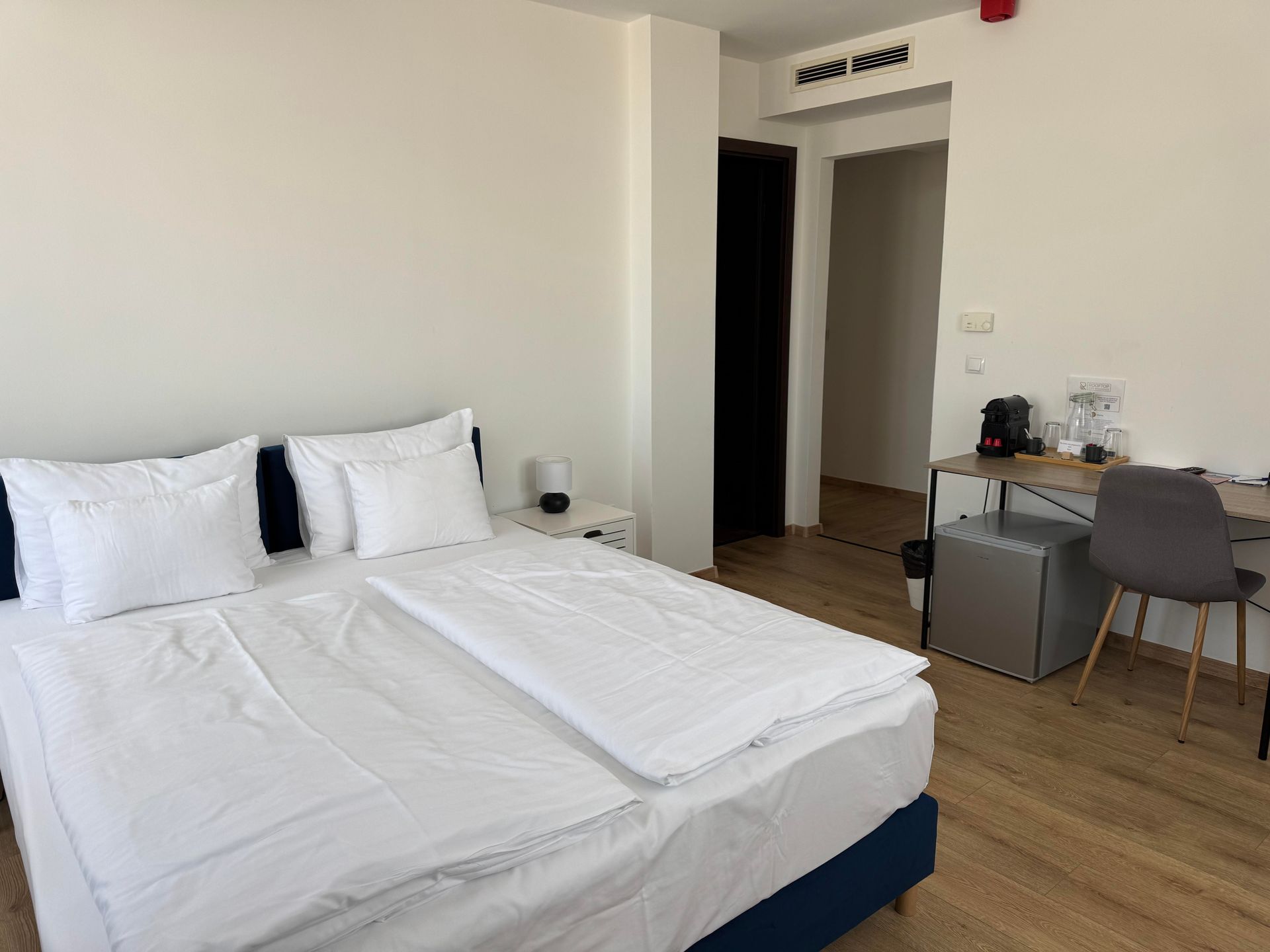 Hotel room with a bed, desk, and a mini-fridge. Walls are white and floors are light wood.