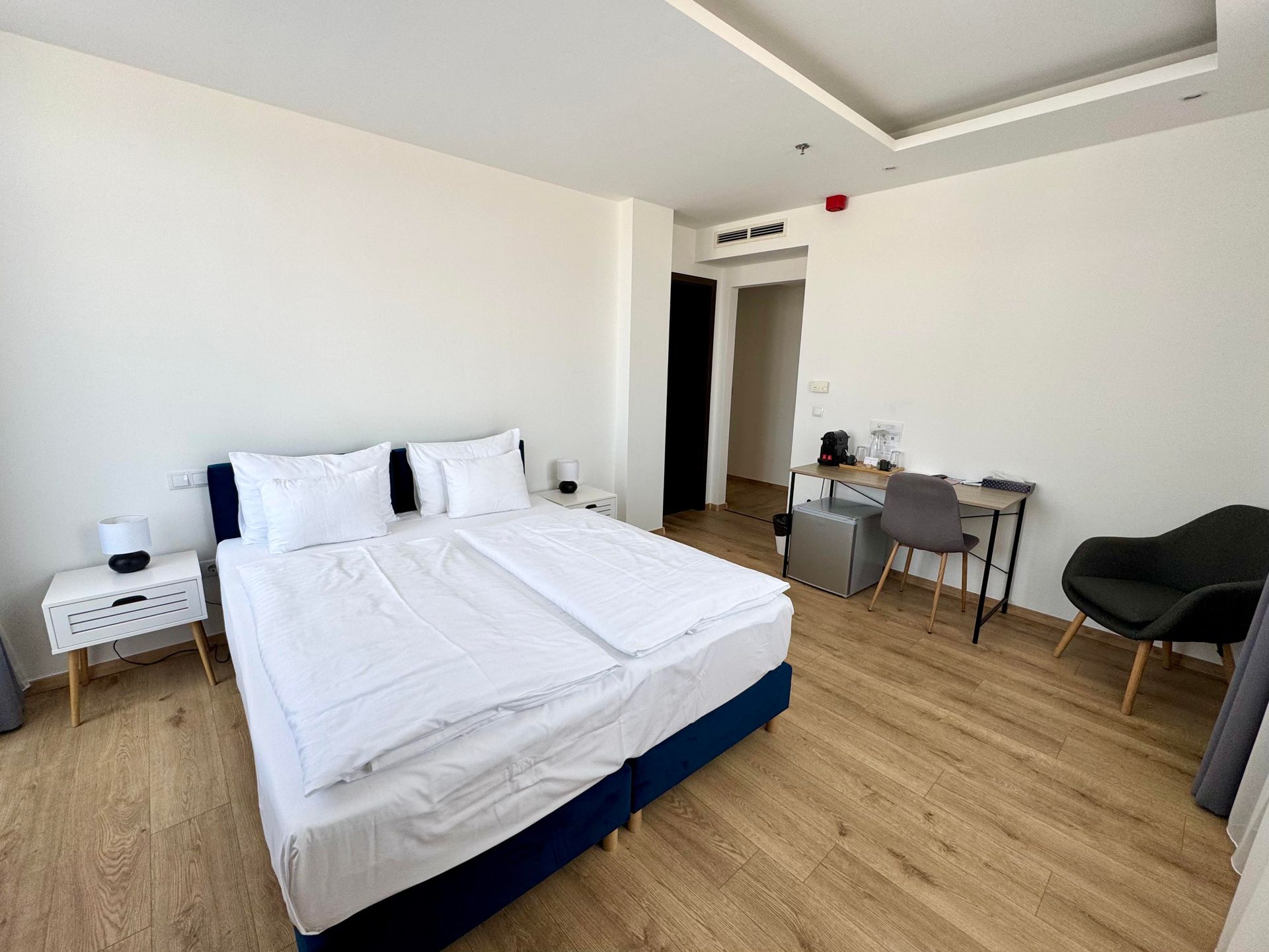 Bedroom with bed, desk, chair, and luggage; light wood floor and white walls.
