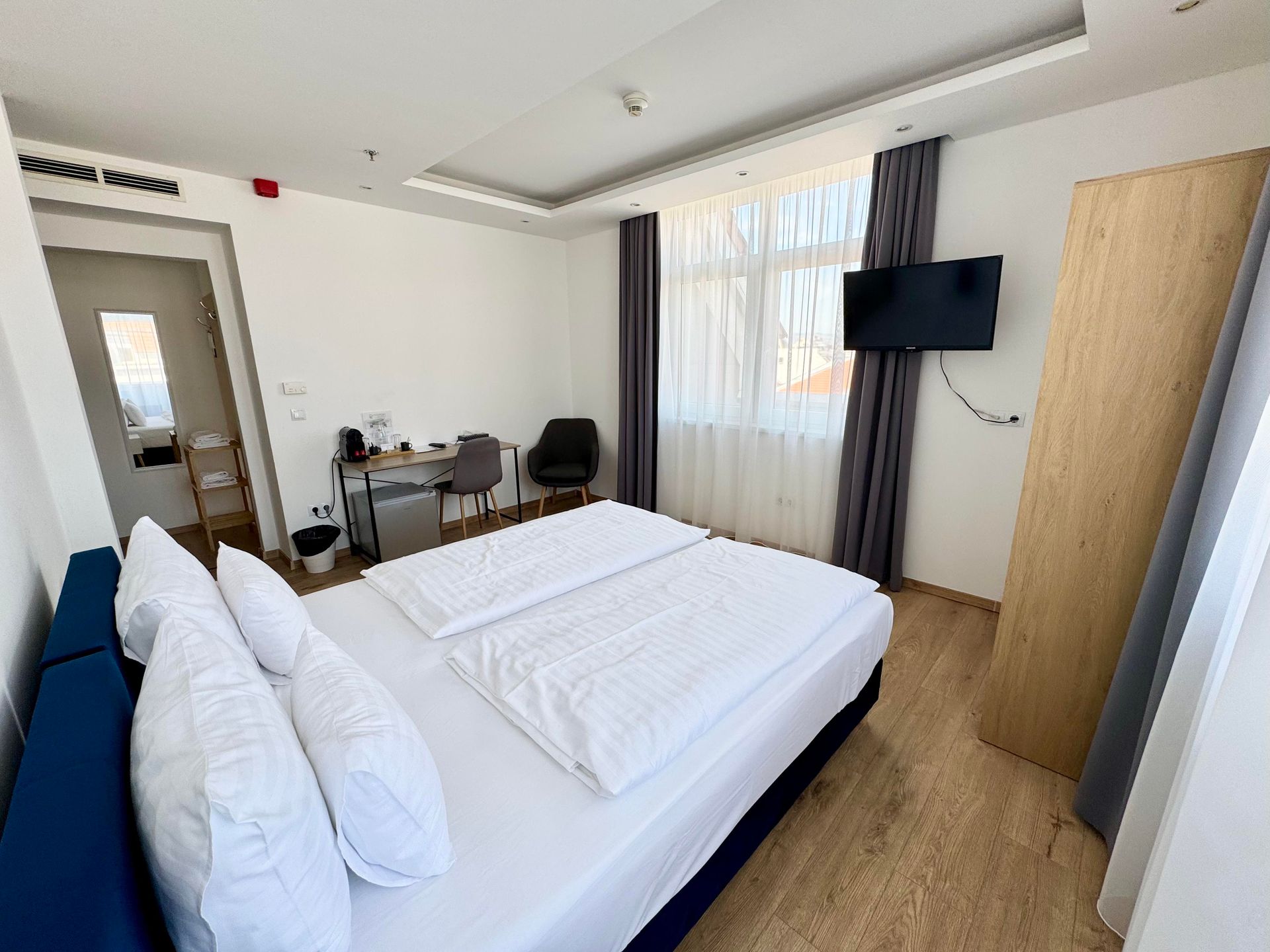 Hotel room with a bed, desk, TV, and window with curtains; light colors and wood floors.