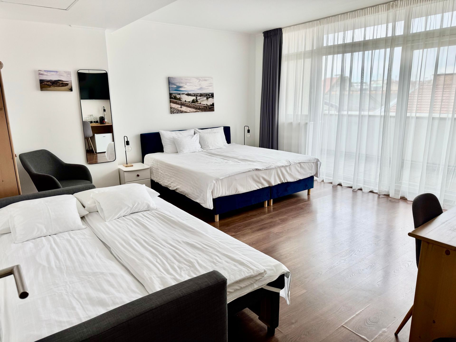 Bedroom with two beds, large windows, and a desk. White bedding, blue bed frames, and dark curtains.