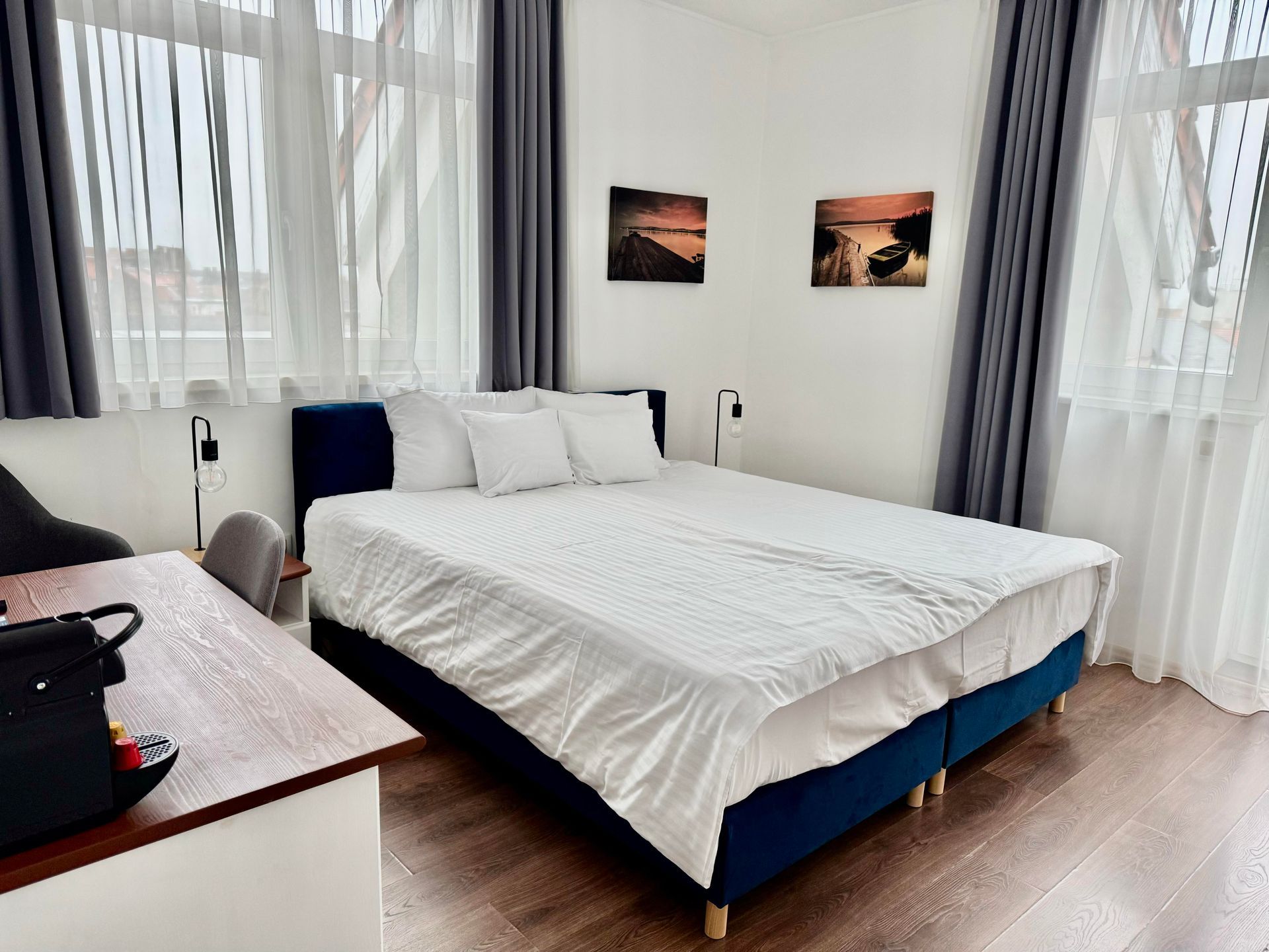 Bedroom with a double bed, desk, and windows with curtains. Two landscape paintings on the wall.