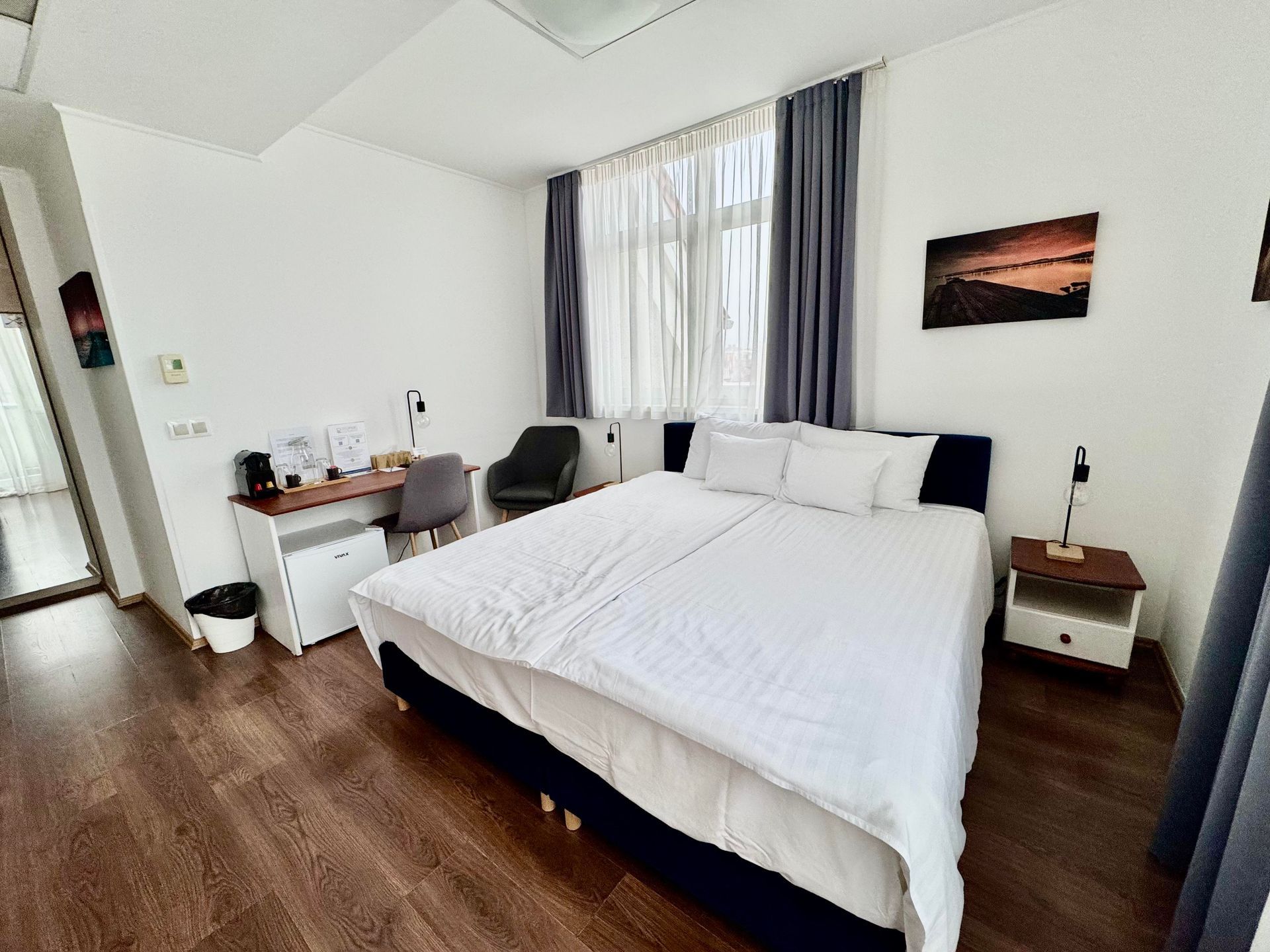 Bedroom with a large bed, desk, and a window with sheer curtains. Wooden floors, white walls.
