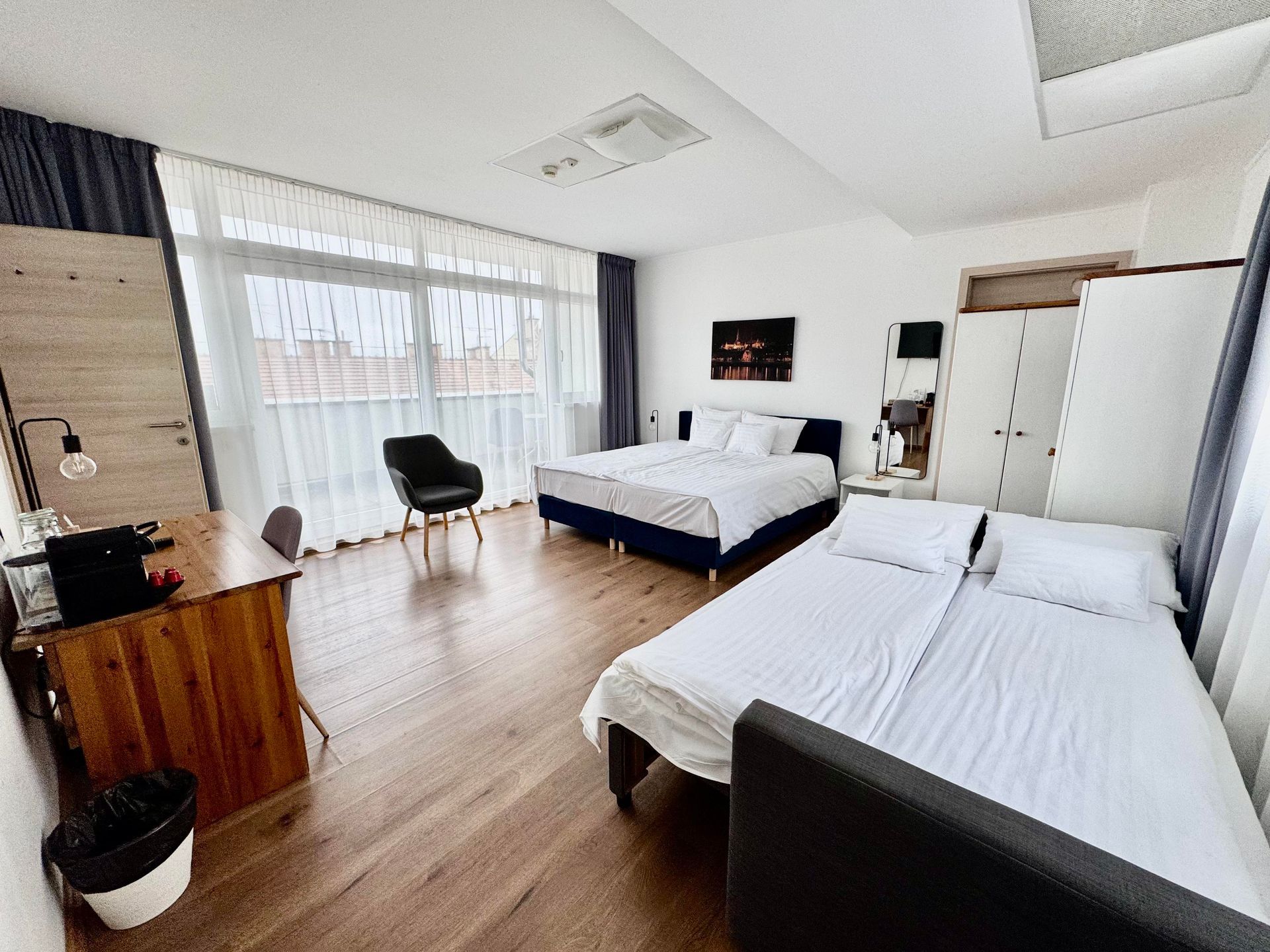 Hotel room with two beds, a desk, and a wardrobe. White walls, dark blue bed frames and curtains.