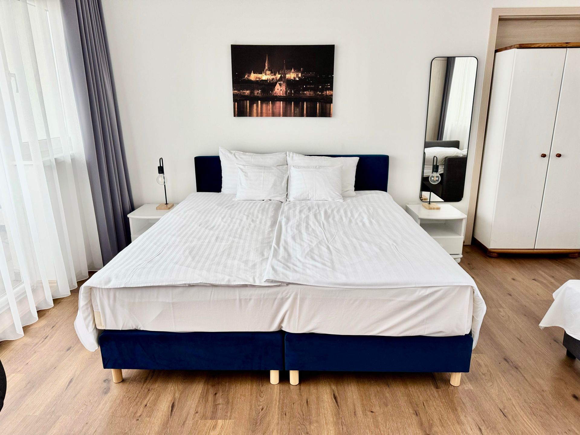 Bedroom with a blue bed frame, white bedding, and a large mirror. A cityscape painting hangs above the bed.