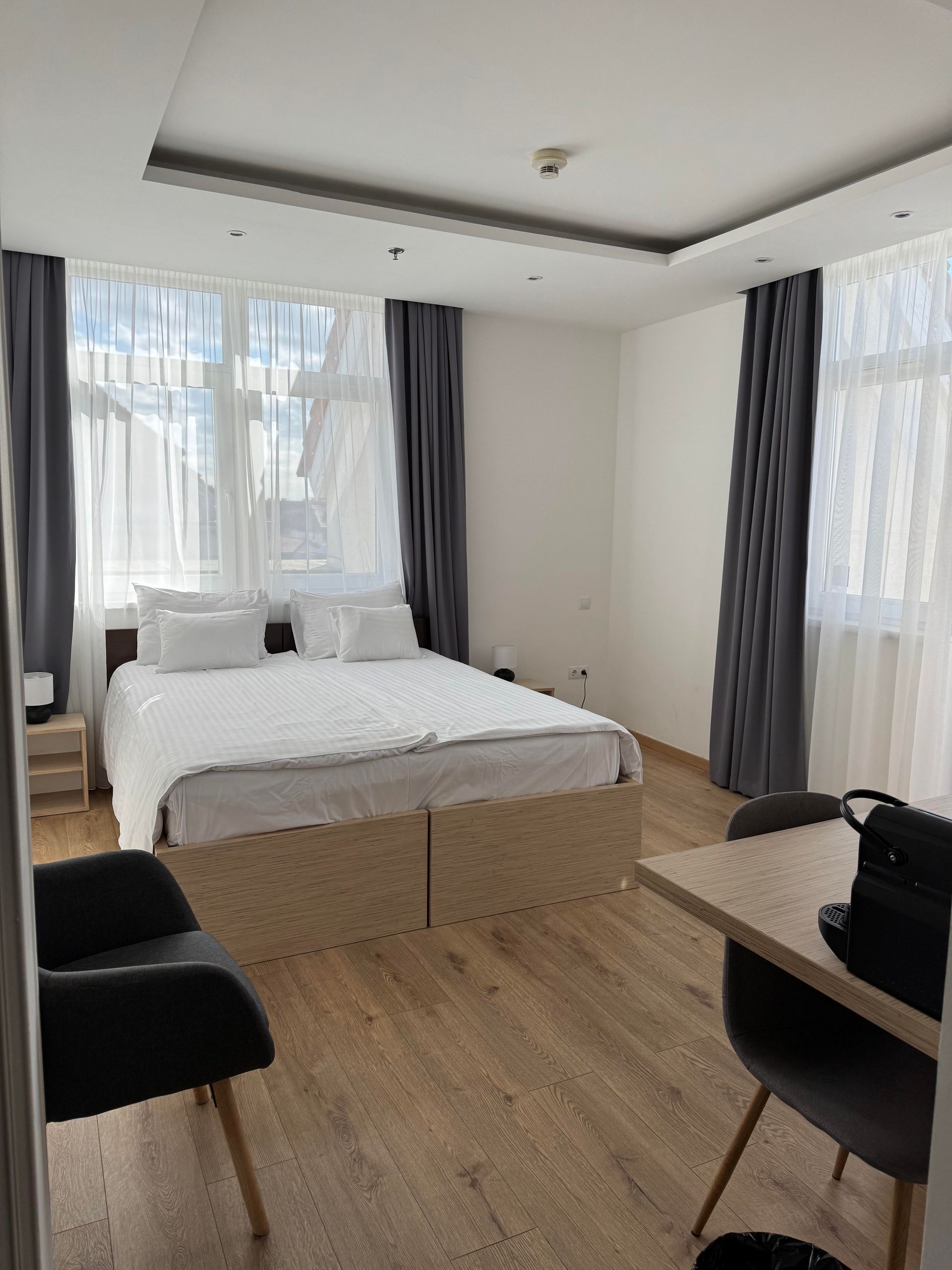 Hotel room with a bed, desk, and chairs near a window with curtains; light wood floors.