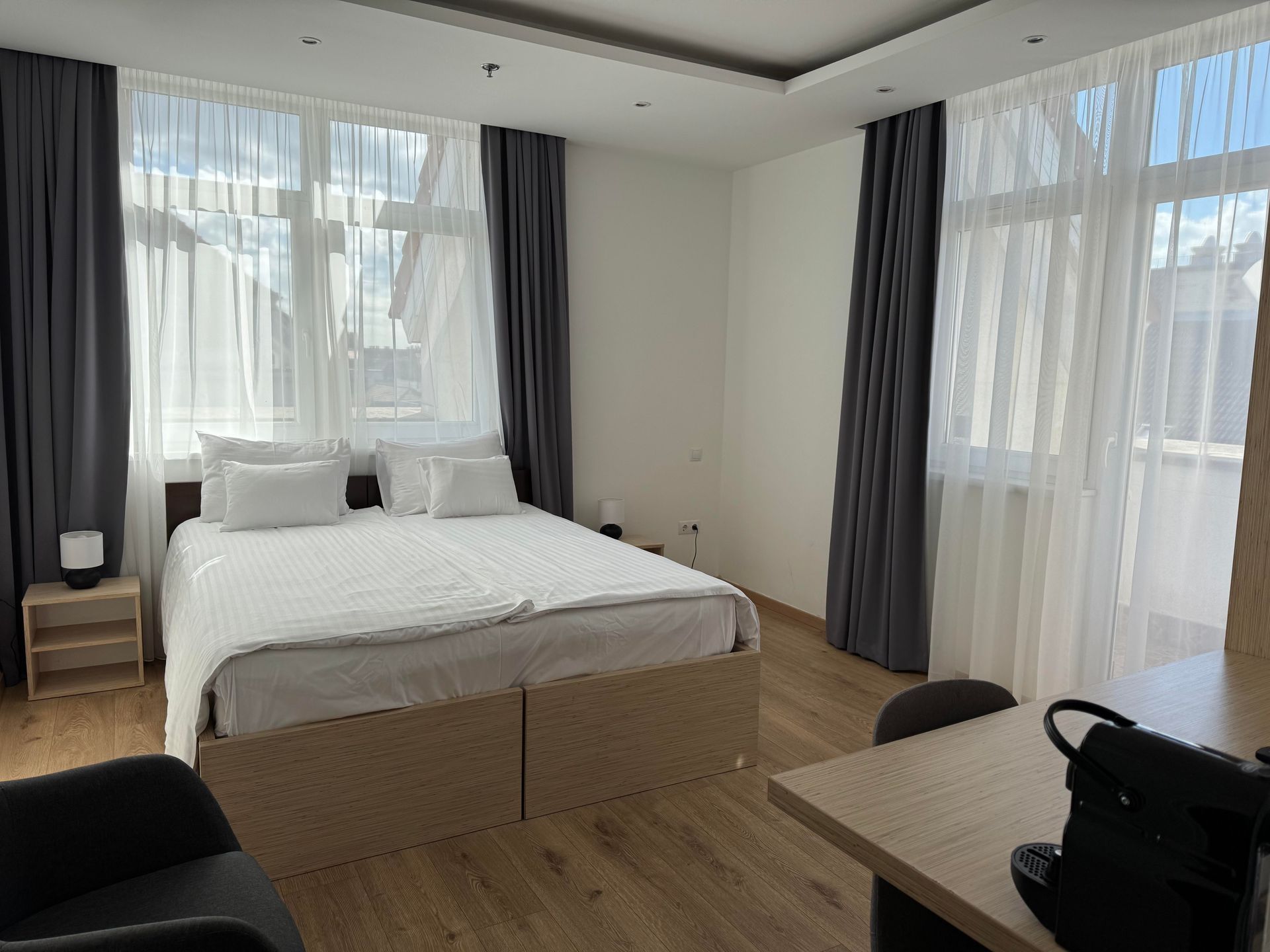 Bedroom with a large bed, sheer curtains, and dark grey drapes.  A small table and chair are in the foreground.