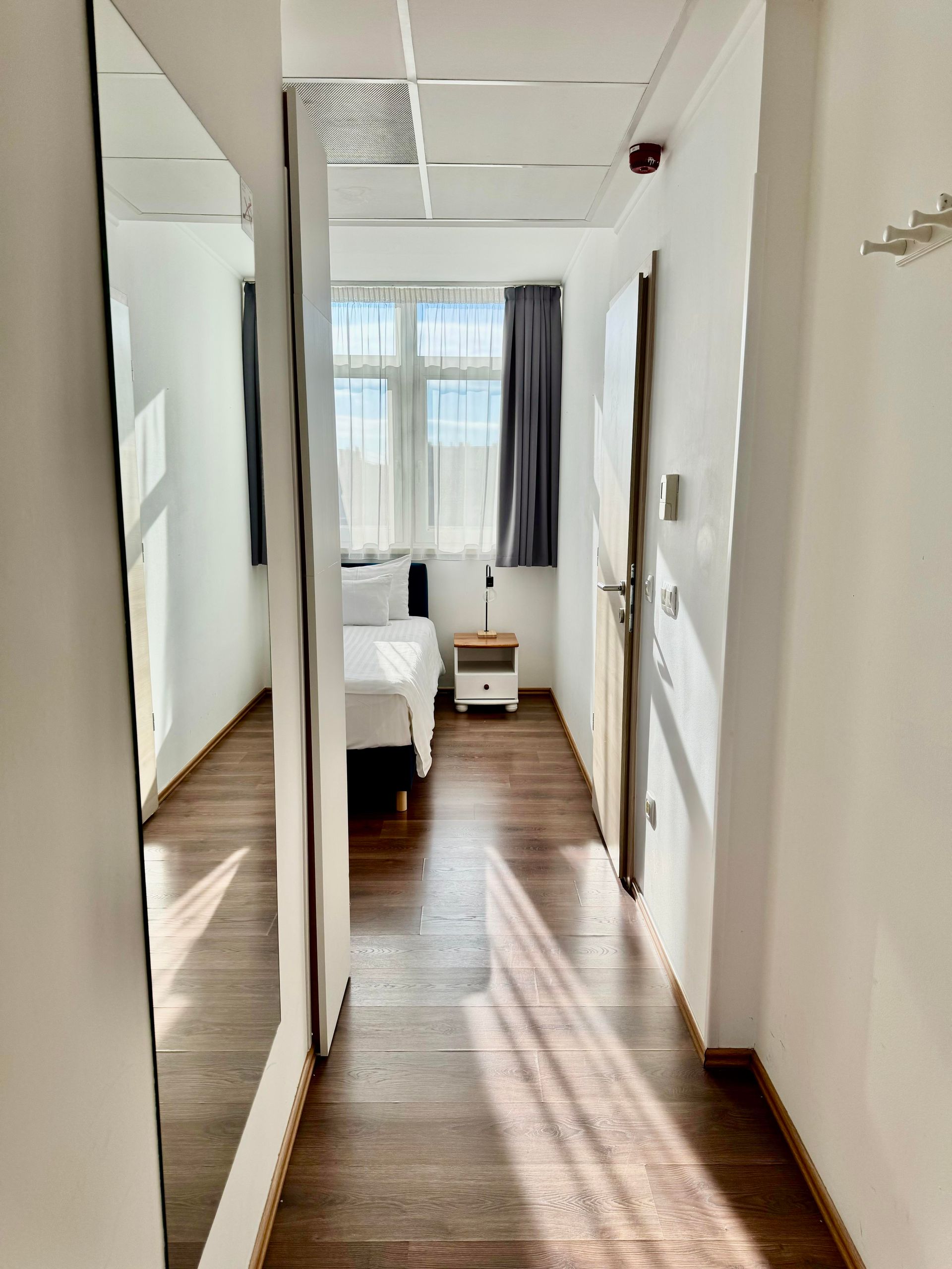 Narrow hallway leading to a bedroom with a bed. Large mirror on the left, wood floor.