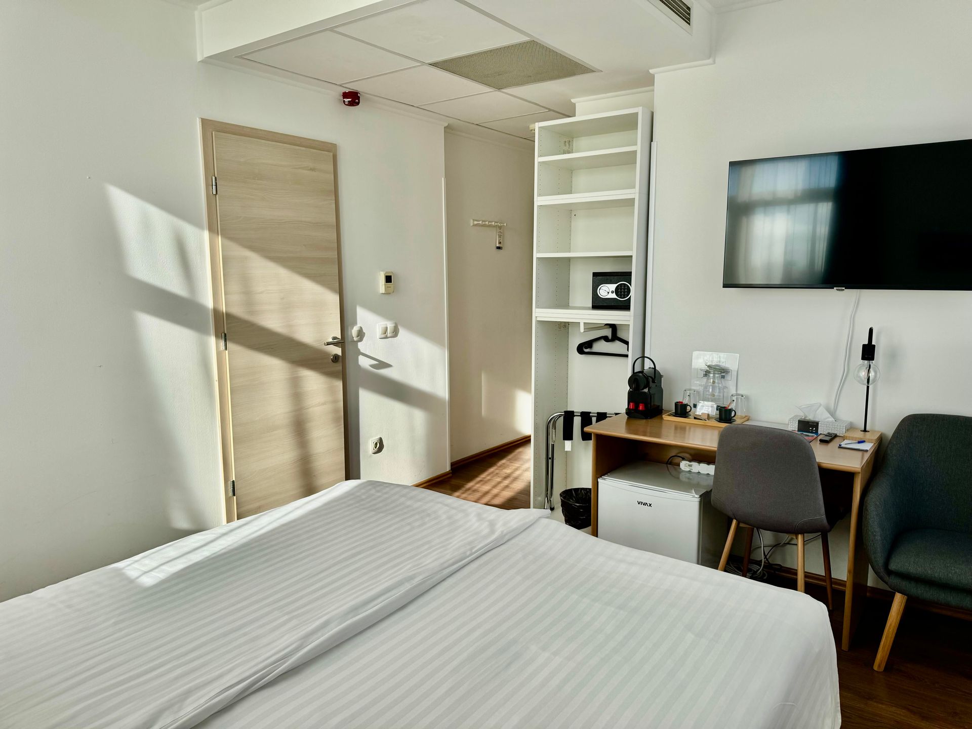 Hotel room with a bed, desk, shelving, TV, and closed door.