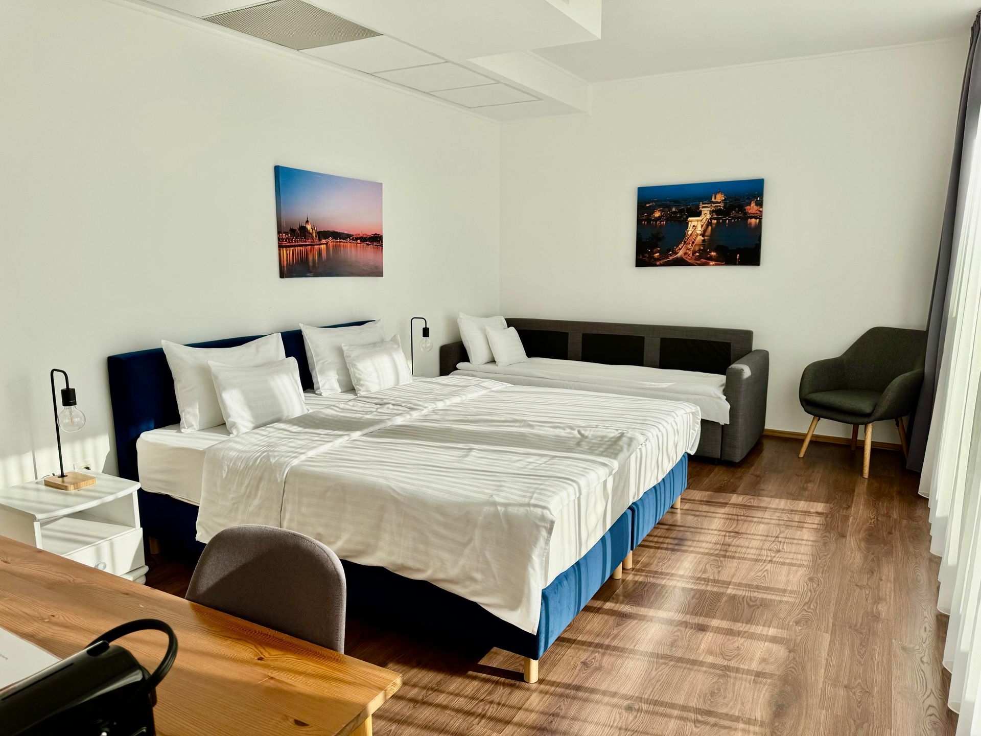 Bright hotel room with a large bed, pull-out sofa, armchair, and city art prints.