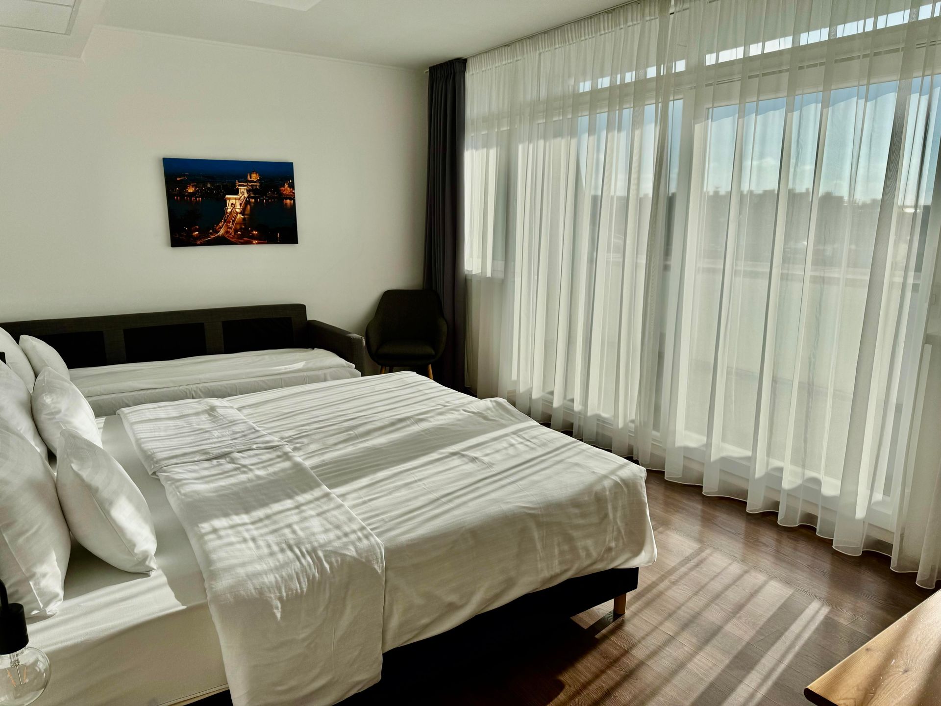 Bedroom with a double bed, single bed, sheer curtains, dark wood floor, and a photo on the wall.