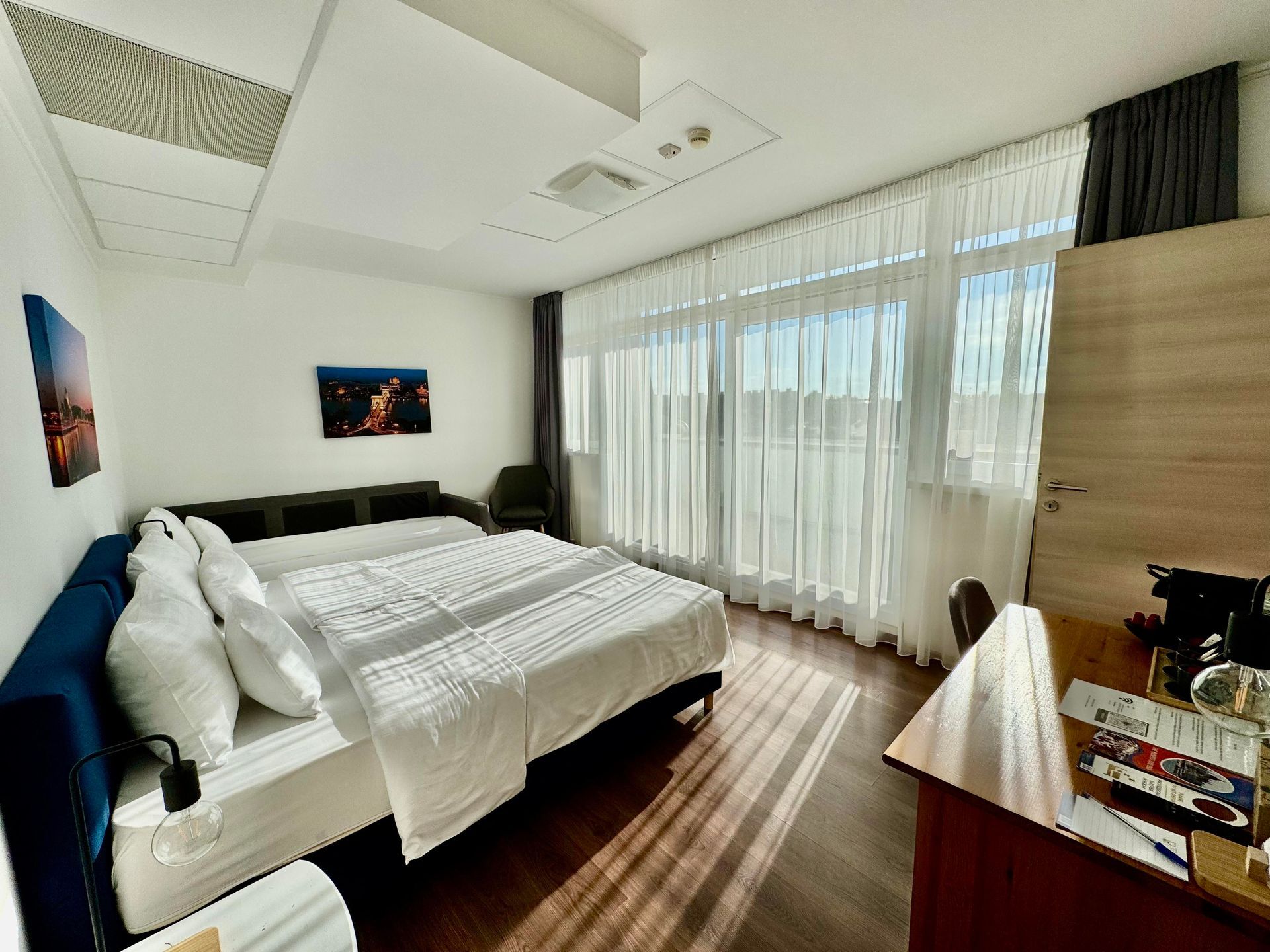 Bedroom with bed, desk, and large window with sheer curtains. Sunlight streams in.