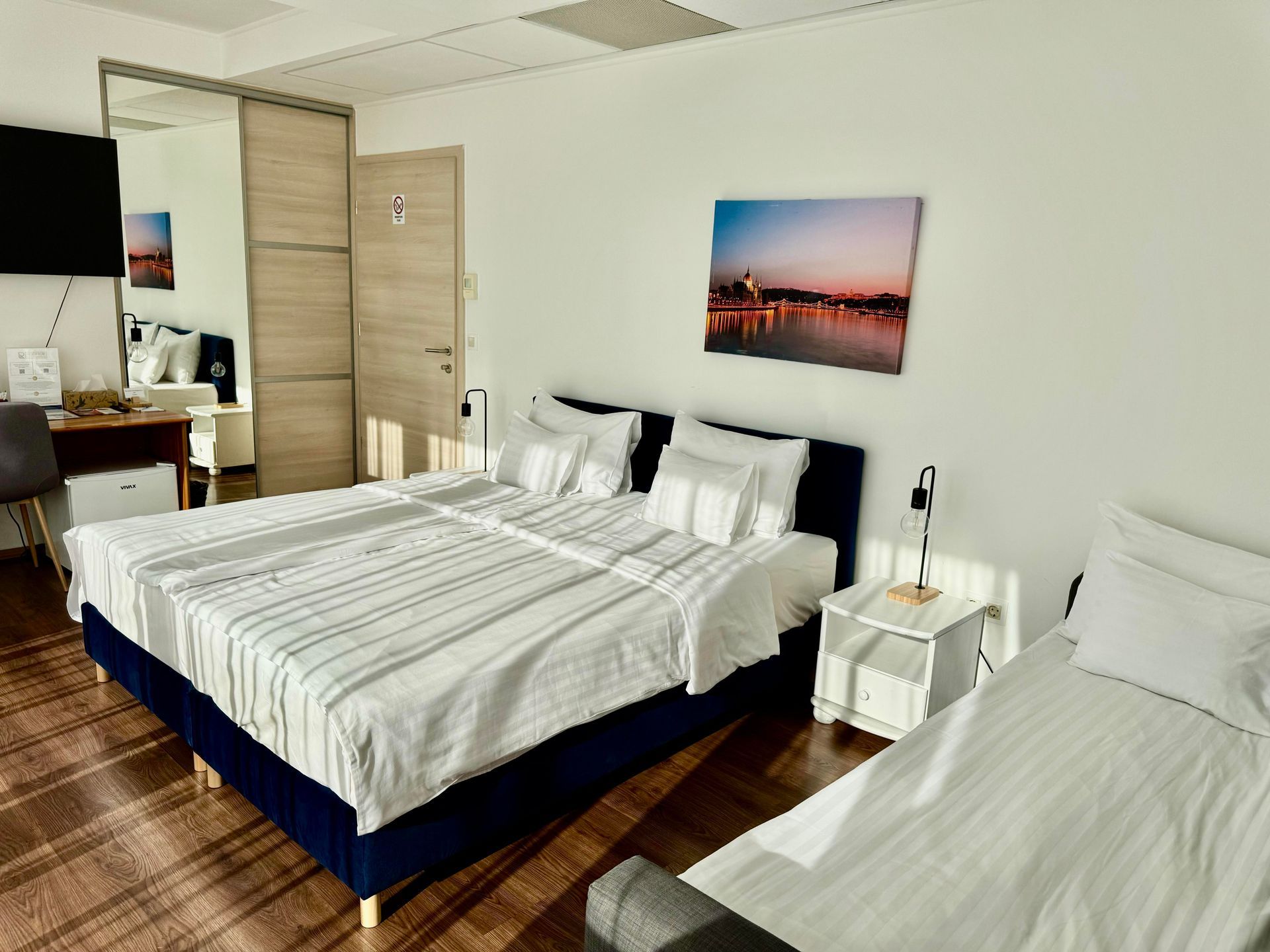 Bright hotel room with two beds, artwork, mirror, and side table.