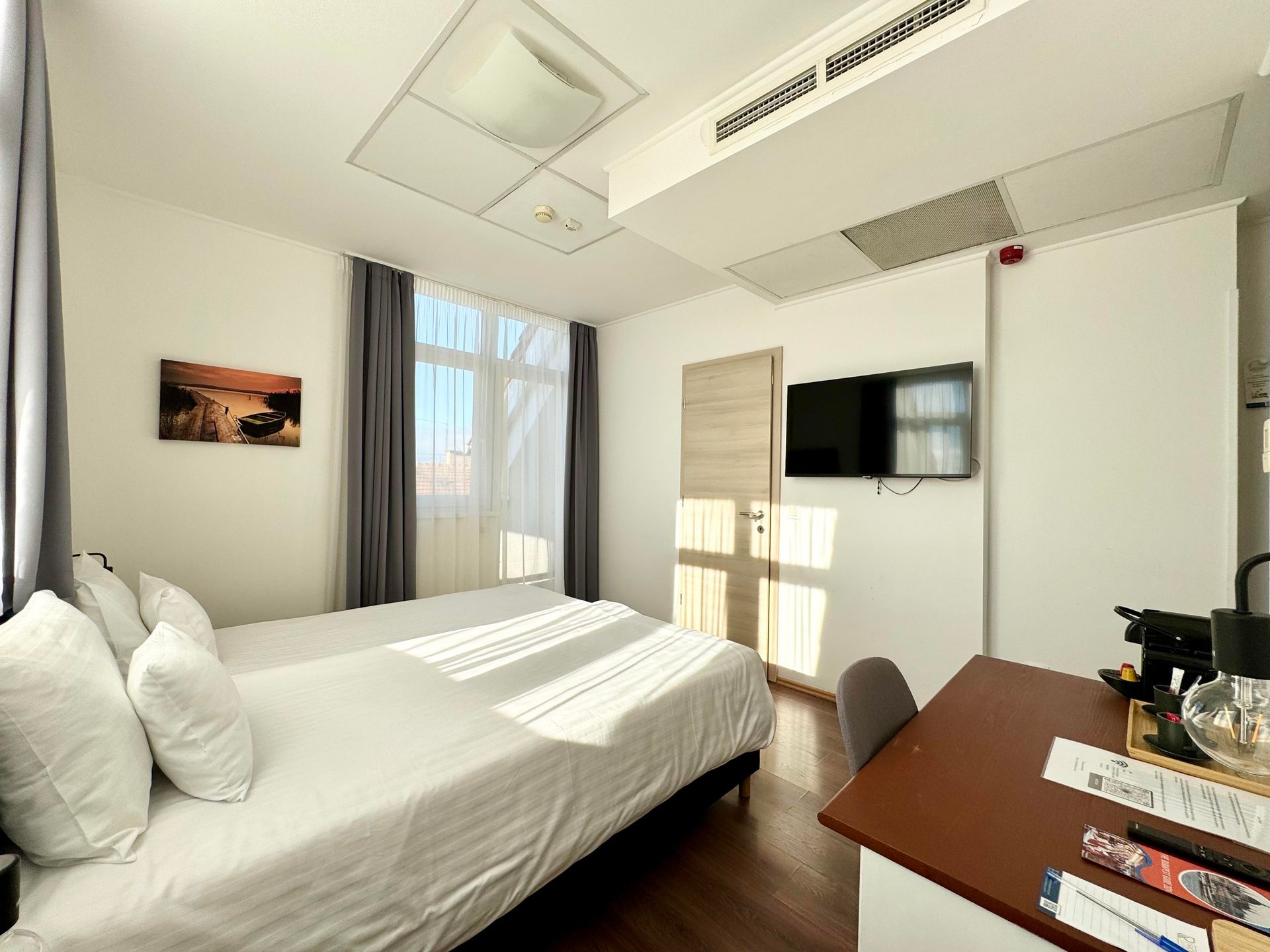 Hotel room with two beds, TV, desk, and sunlight through the window.