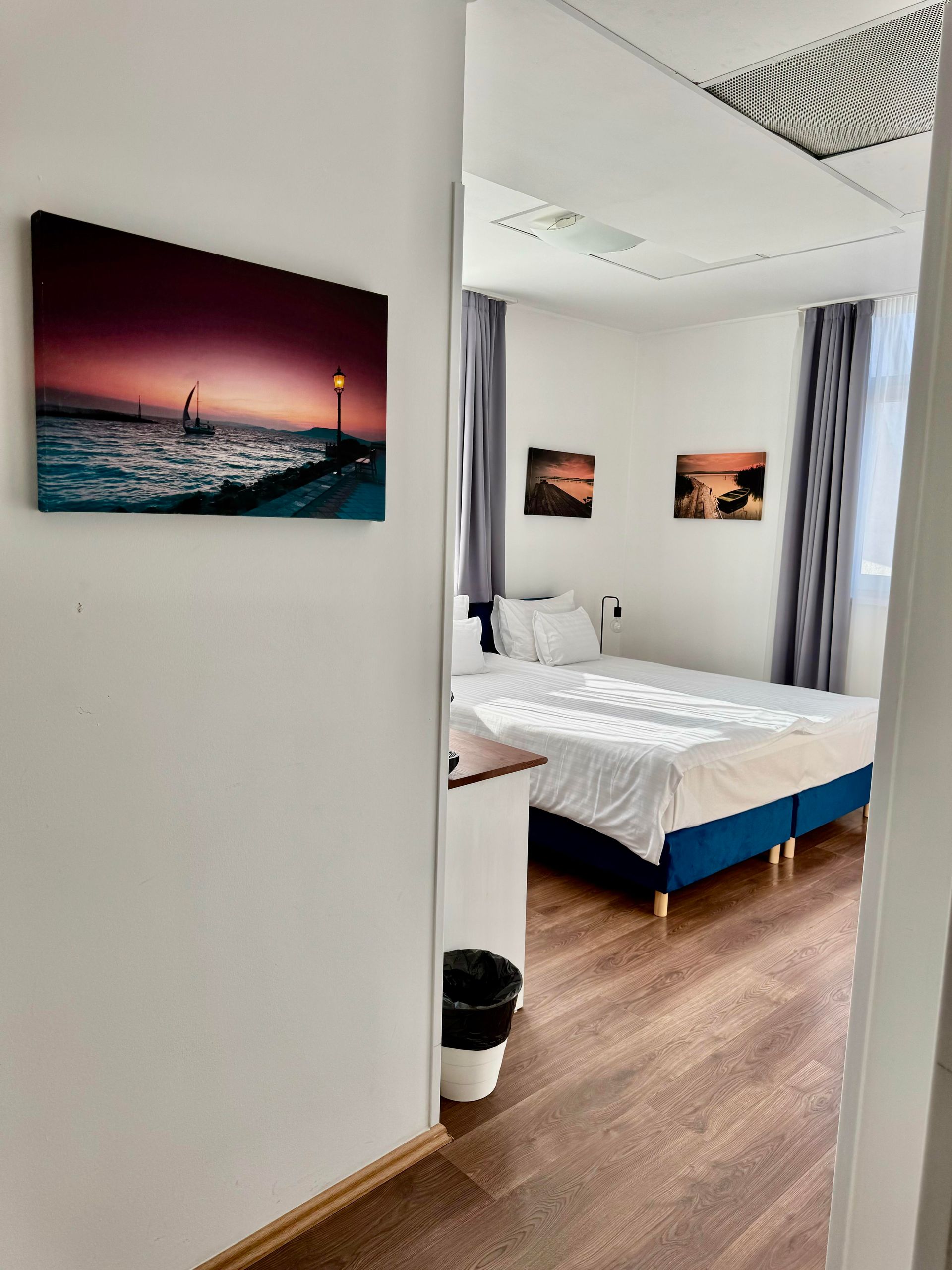 Bedroom interior, view from doorway. Bed with white linens, artwork on walls, wood floor, and dark curtains.