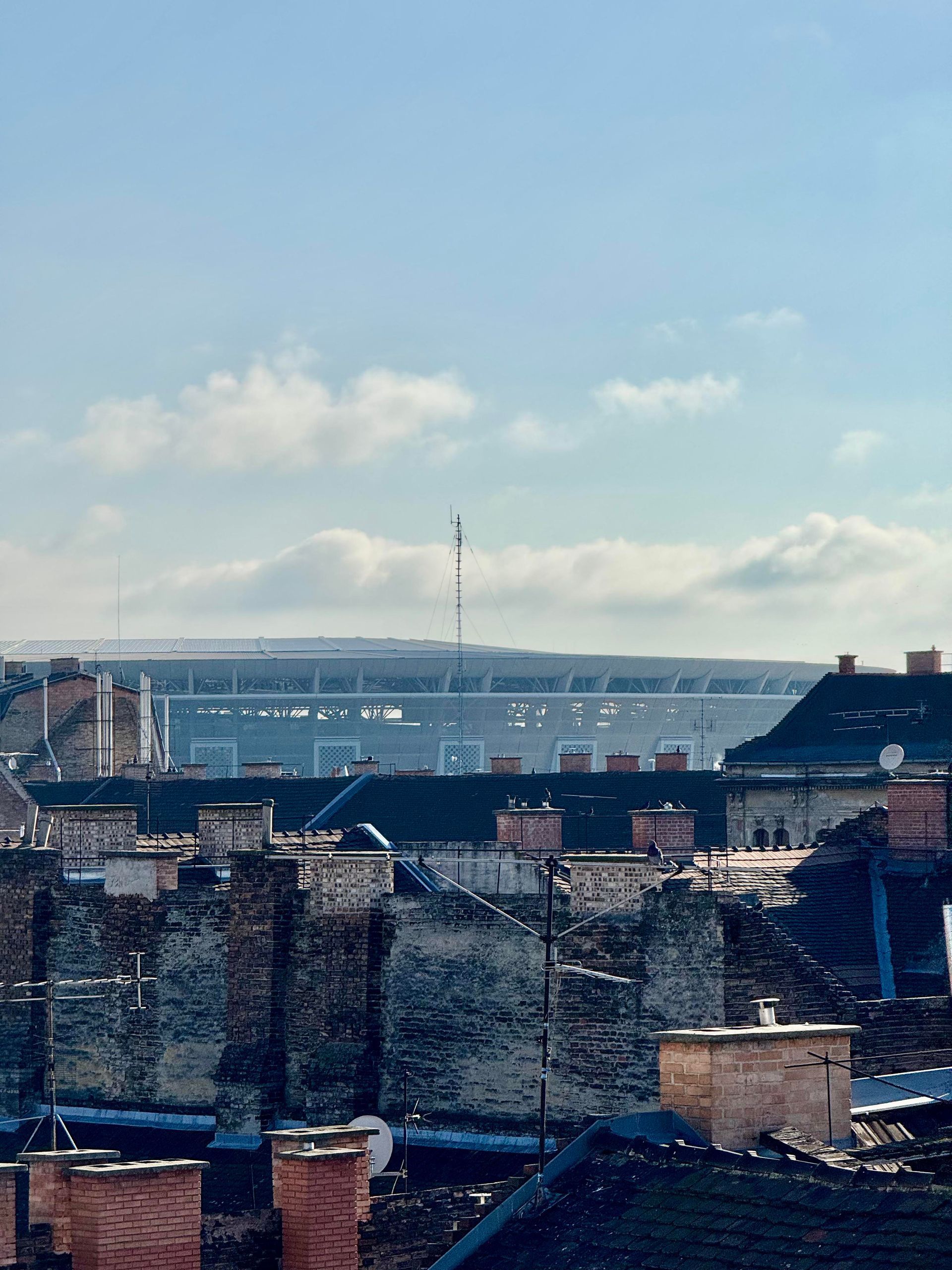 Rooftops with brick chimneys overlook a distant stadium on a sunny day with a blue sky.