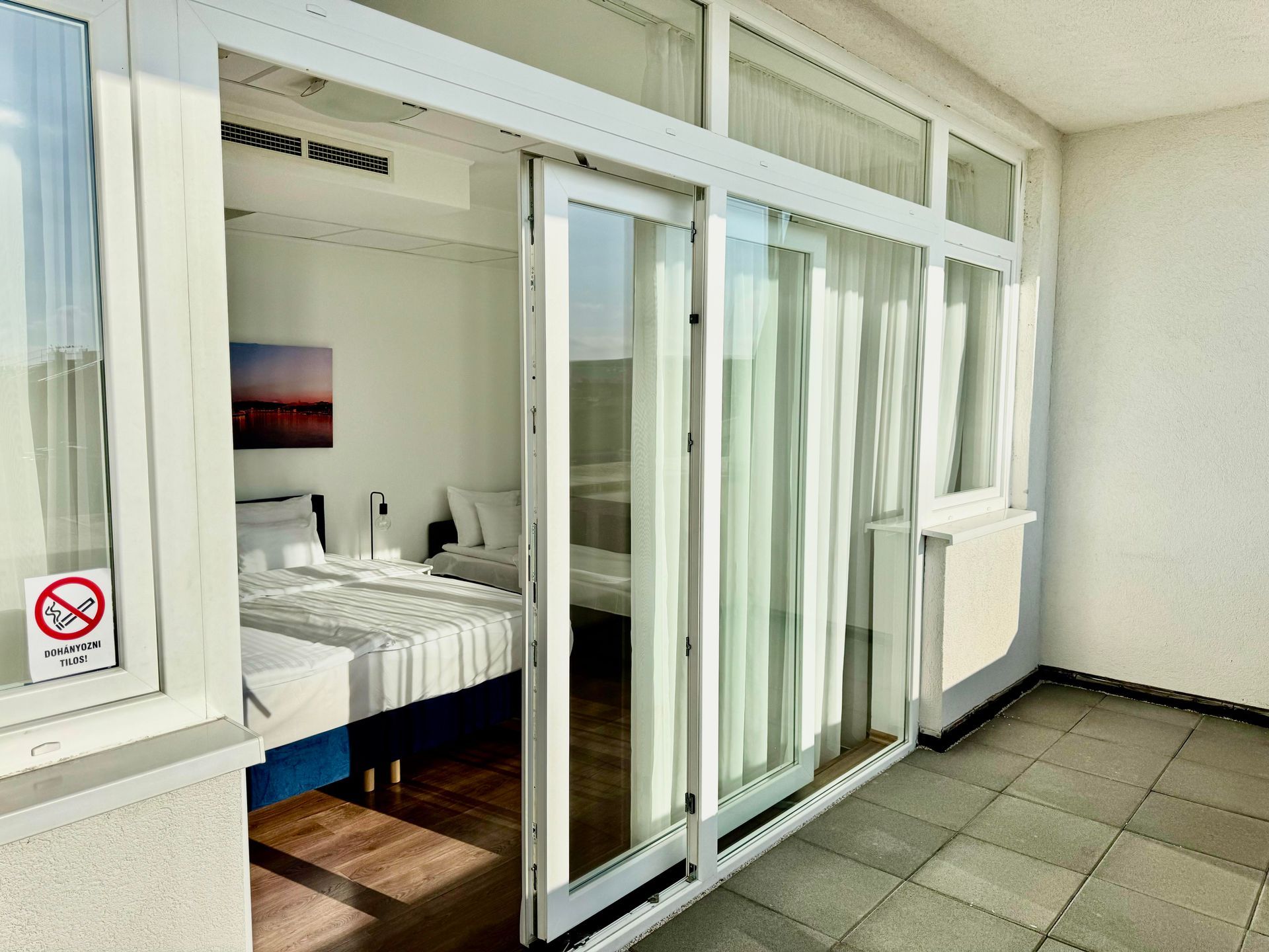 Sliding glass doors open from a balcony to a bright white room with a bed; a no smoking sign is visible.