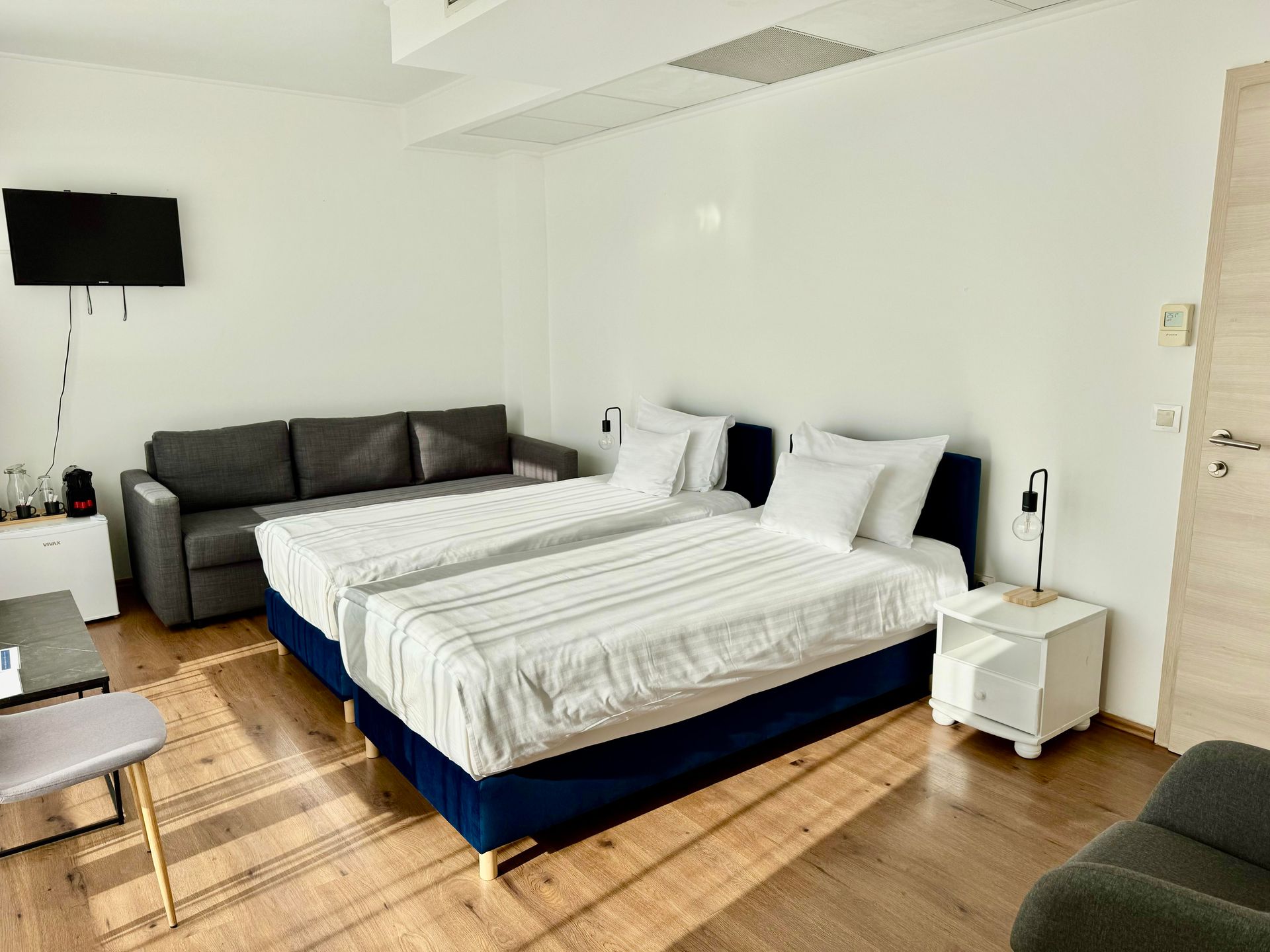 Hotel room with a bed, sofa, TV, and side tables; wooden floors and white walls.