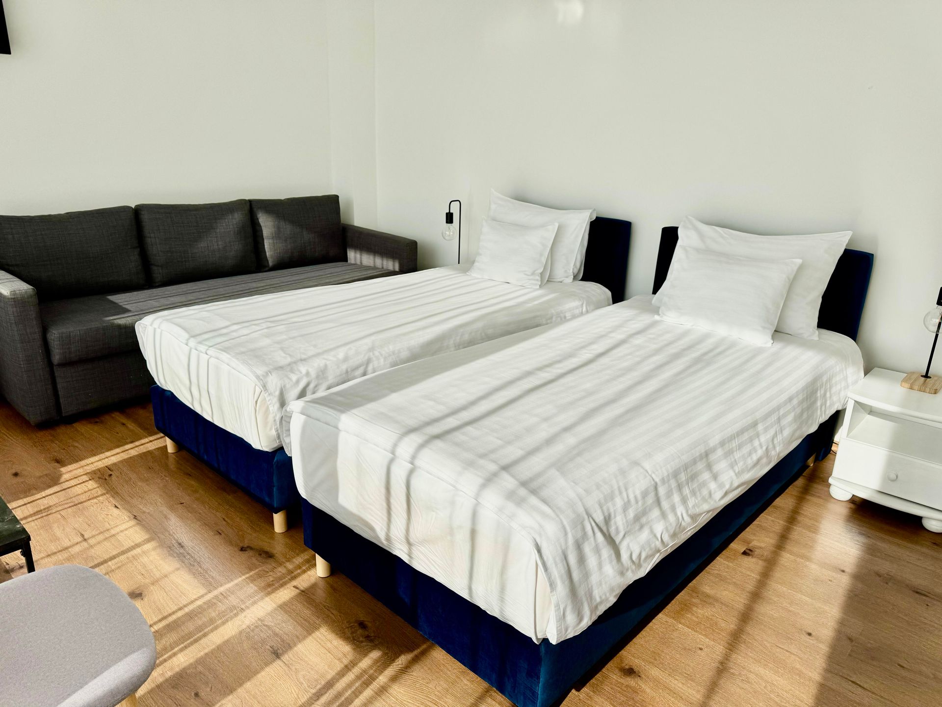 Two twin beds with white bedding, a gray couch, and white bedside table in a room with wood flooring.