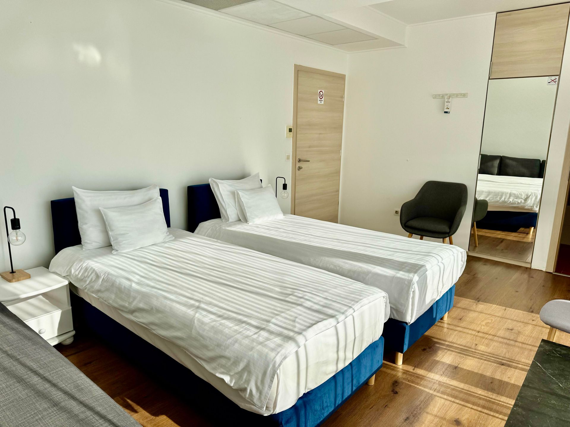 A hotel room with two twin beds with white linens, a wooden floor, and a full-length mirror.