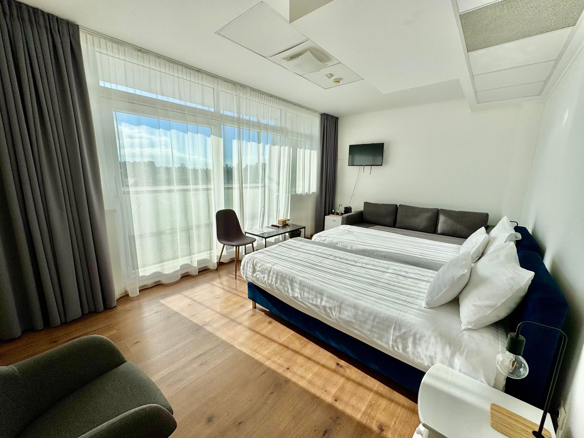 Bright hotel room with two beds, sofa, and floor-to-ceiling windows with sheer curtains. Sunlight streams in.
