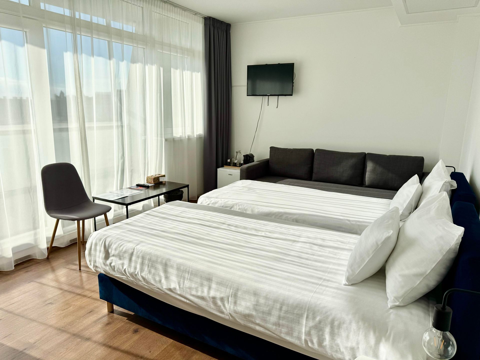 Bedroom with two beds, sofa, chair, small table, and television. Natural light streams in from large windows.