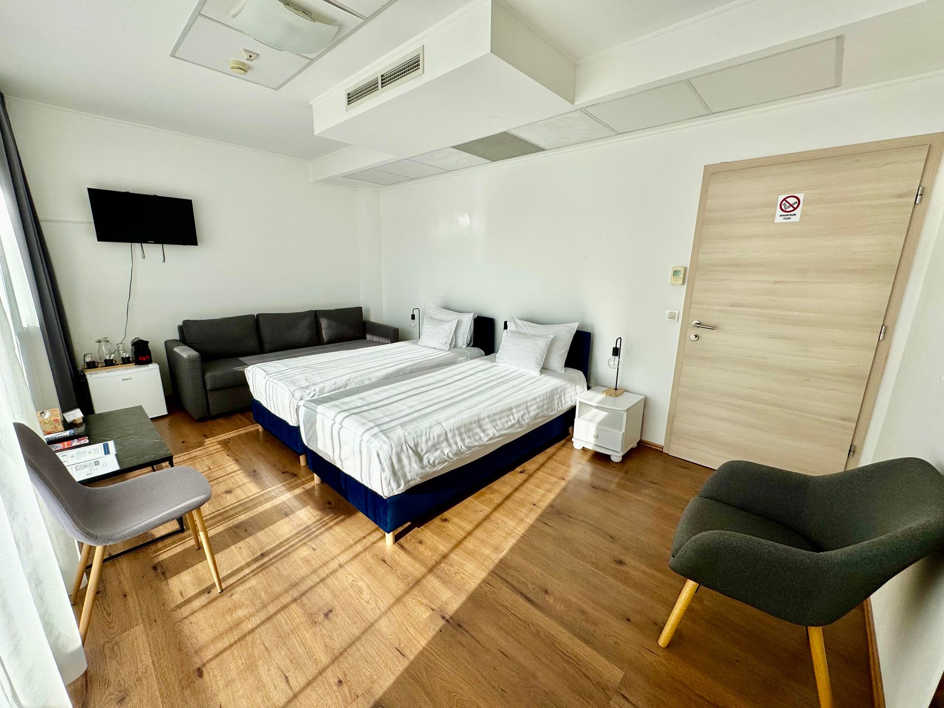 Hotel room with two beds, a sofa, TV, and chair. Wooden floors and natural light.