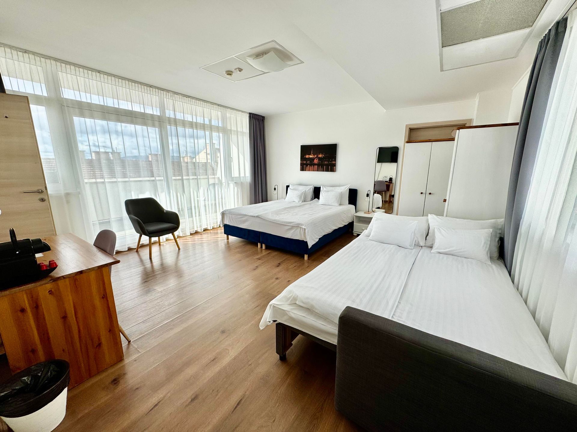 Spacious hotel room with two beds, desk, armchair, and sheer curtains. Bright interior with hardwood floors.