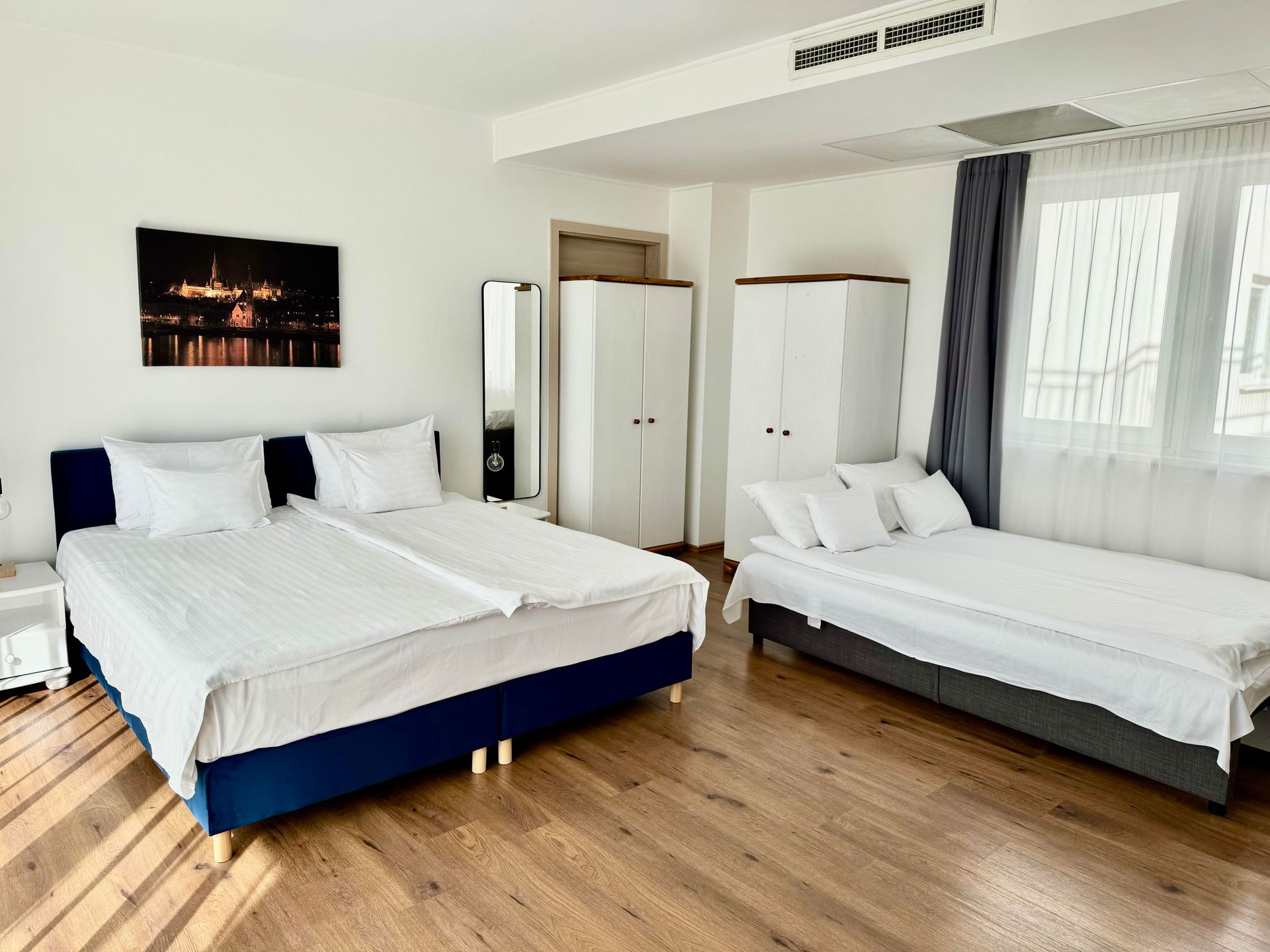 Bedroom with two beds, a single bed, wardrobes, hardwood floor, white walls, and a window.