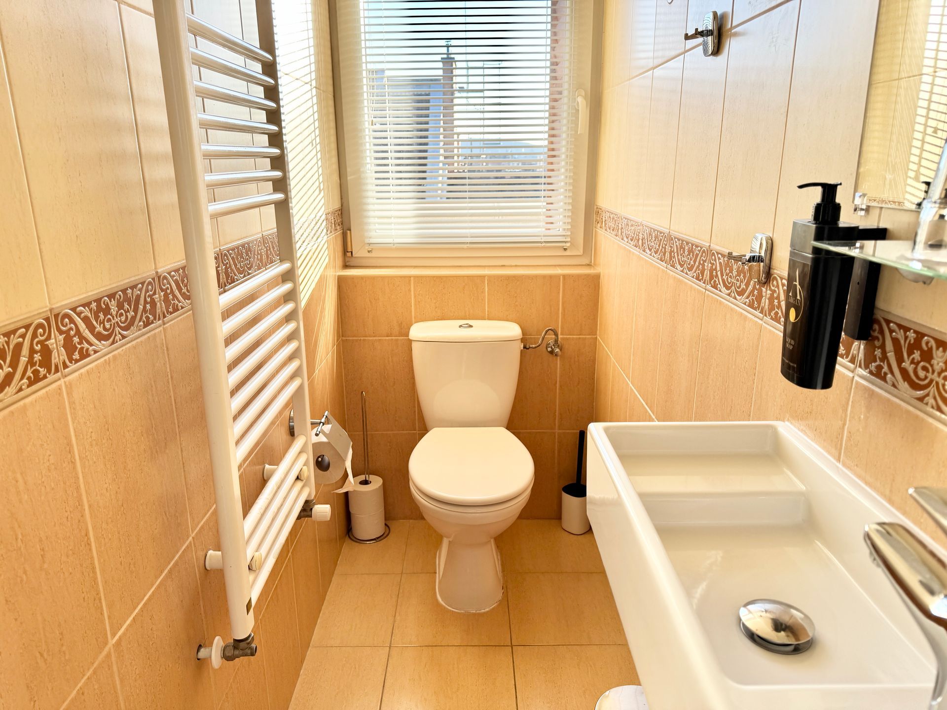 Small bathroom with toilet, sink, and window; tan tiles and white radiator.