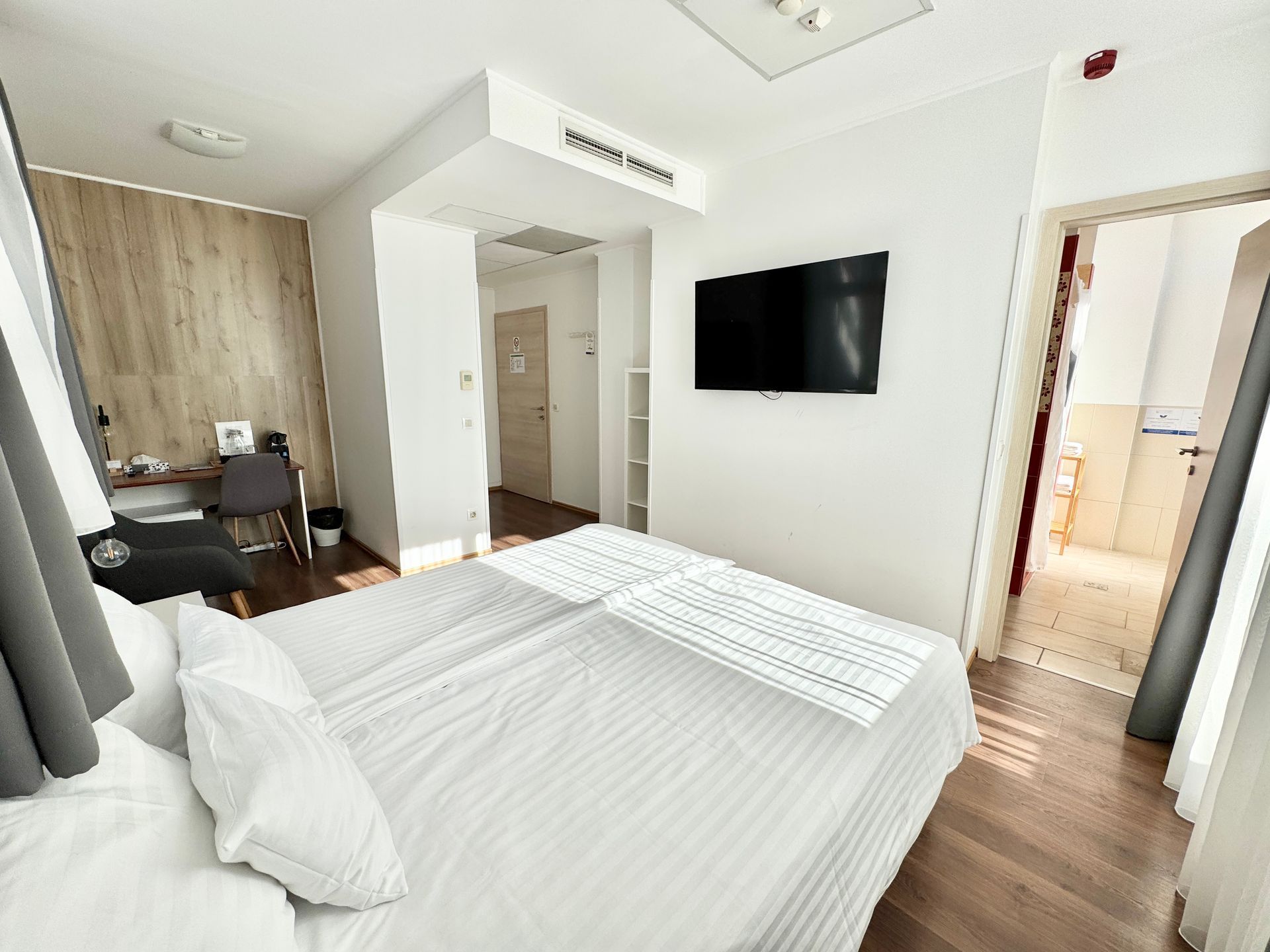 Hotel room with a bed, TV, and open doorway to a bathroom. Wooden floors and accent wall.