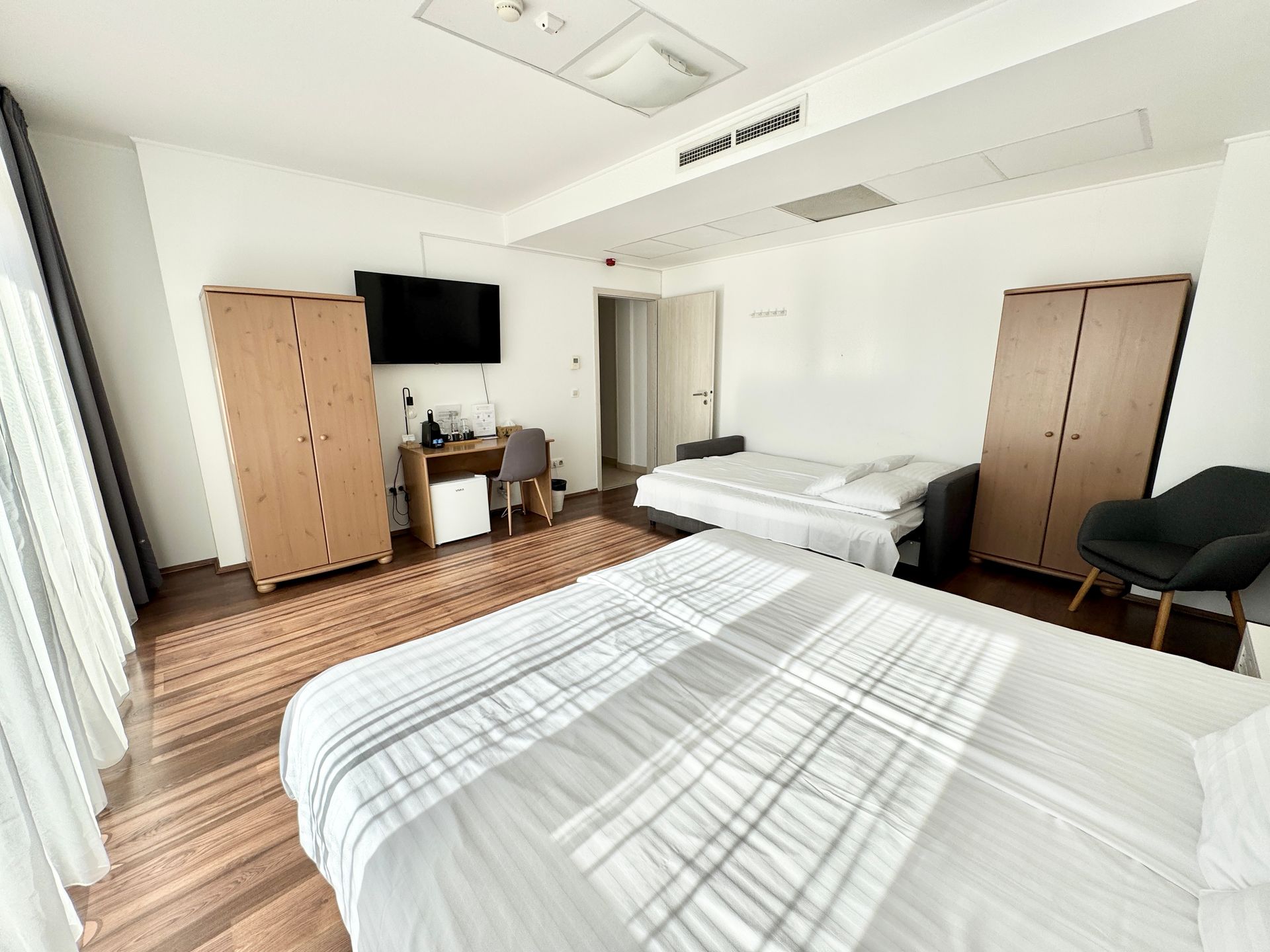 Hotel room with two beds, two wardrobes, a desk, a chair, and a TV. Bright, white walls, wooden floor.