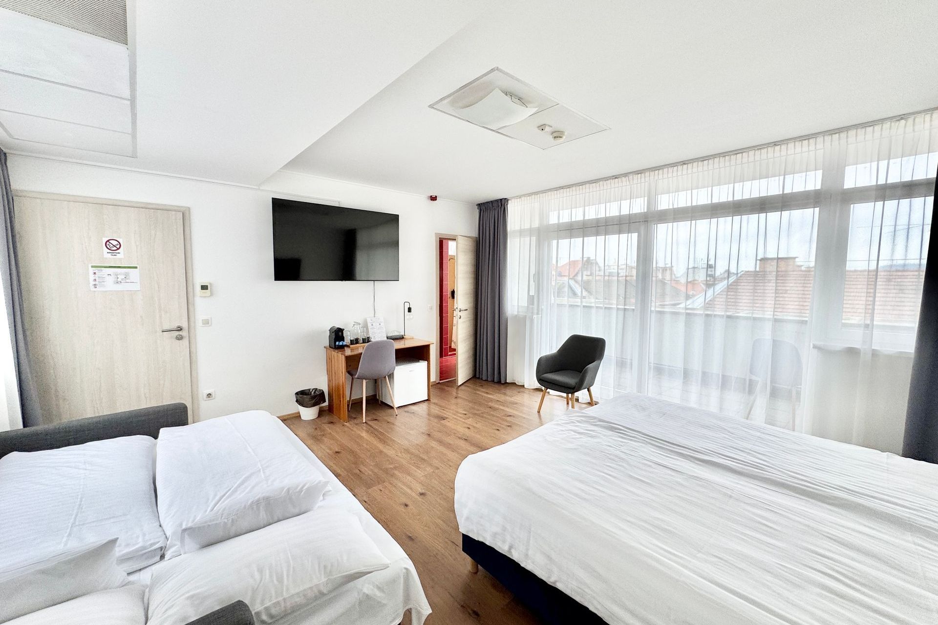 Hotel room with two beds, desk, armchair, TV, and large windows.