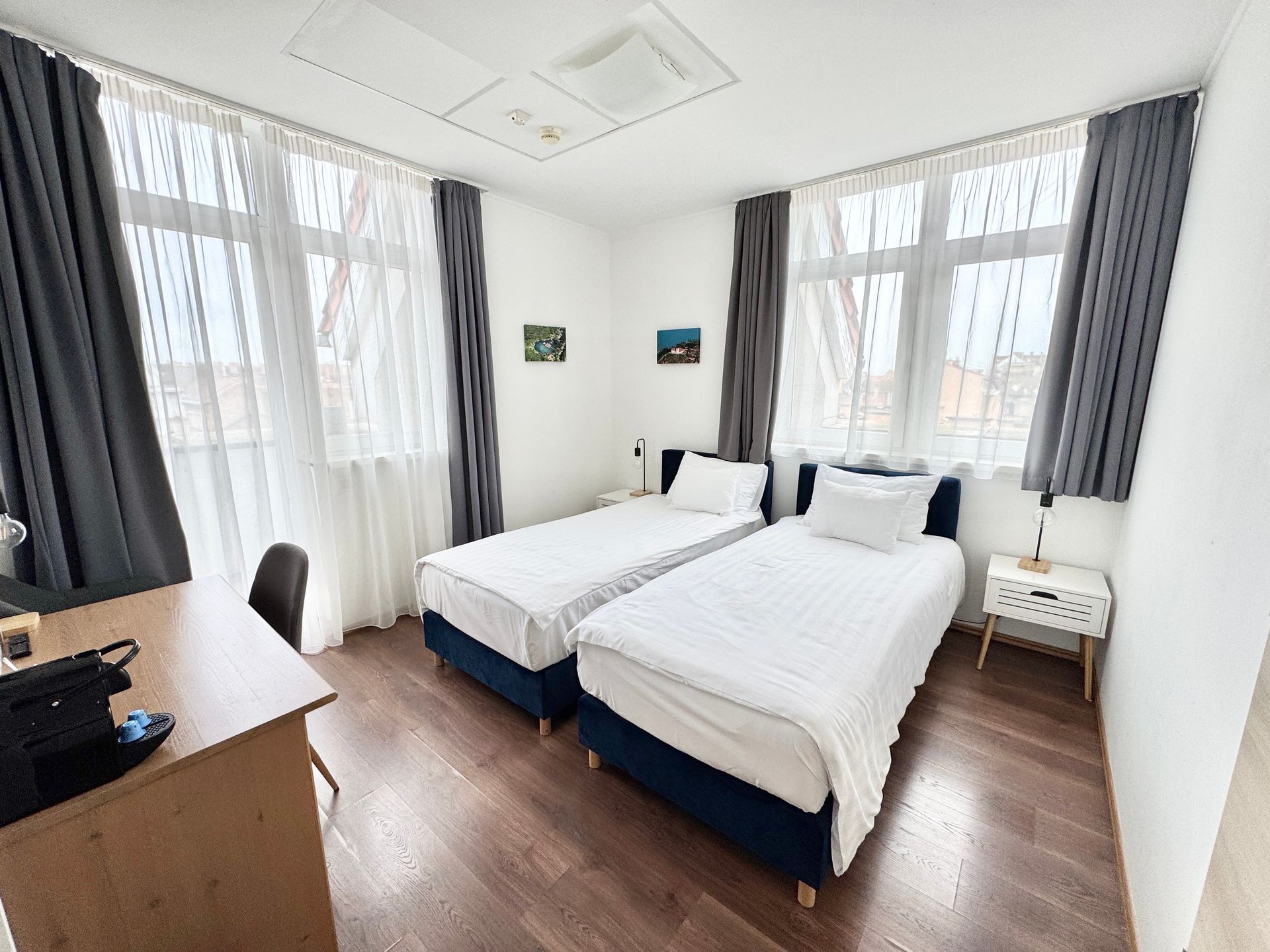 Bedroom with two twin beds, desk, two windows with gray curtains, and a nightstand.