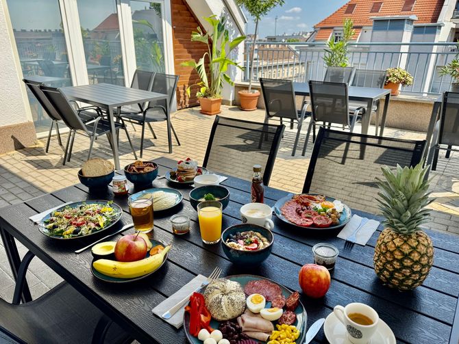 Breakfast spread on a rooftop terrace with tables, chairs, food, drinks, and a city view.