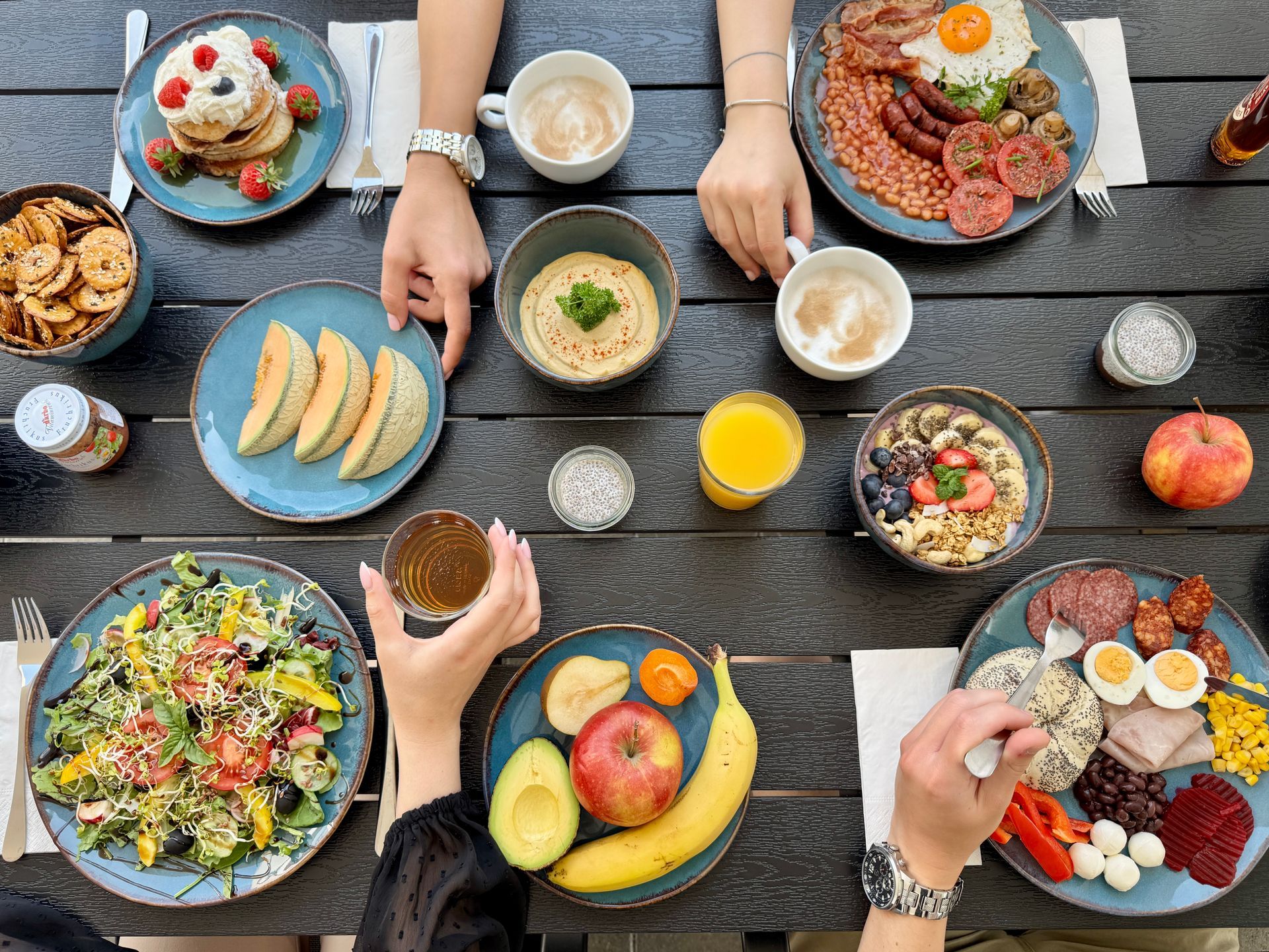 Overhead shot of a table laden with breakfast foods like pancakes, eggs, fruits, and yogurt. Hands reach for the food.