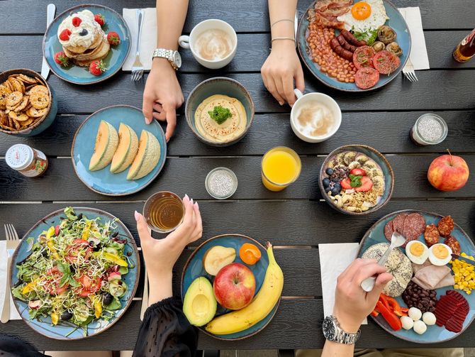 Overhead shot of a table laden with breakfast foods like pancakes, eggs, fruits, and yogurt. Hands reach for the food.