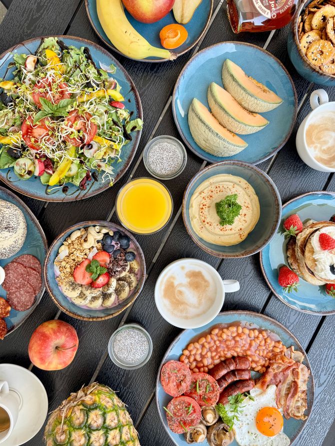 Overhead shot of a brunch spread on a wooden table. Dishes include fruit, salads, oatmeal, pancakes, and a full English breakfast.