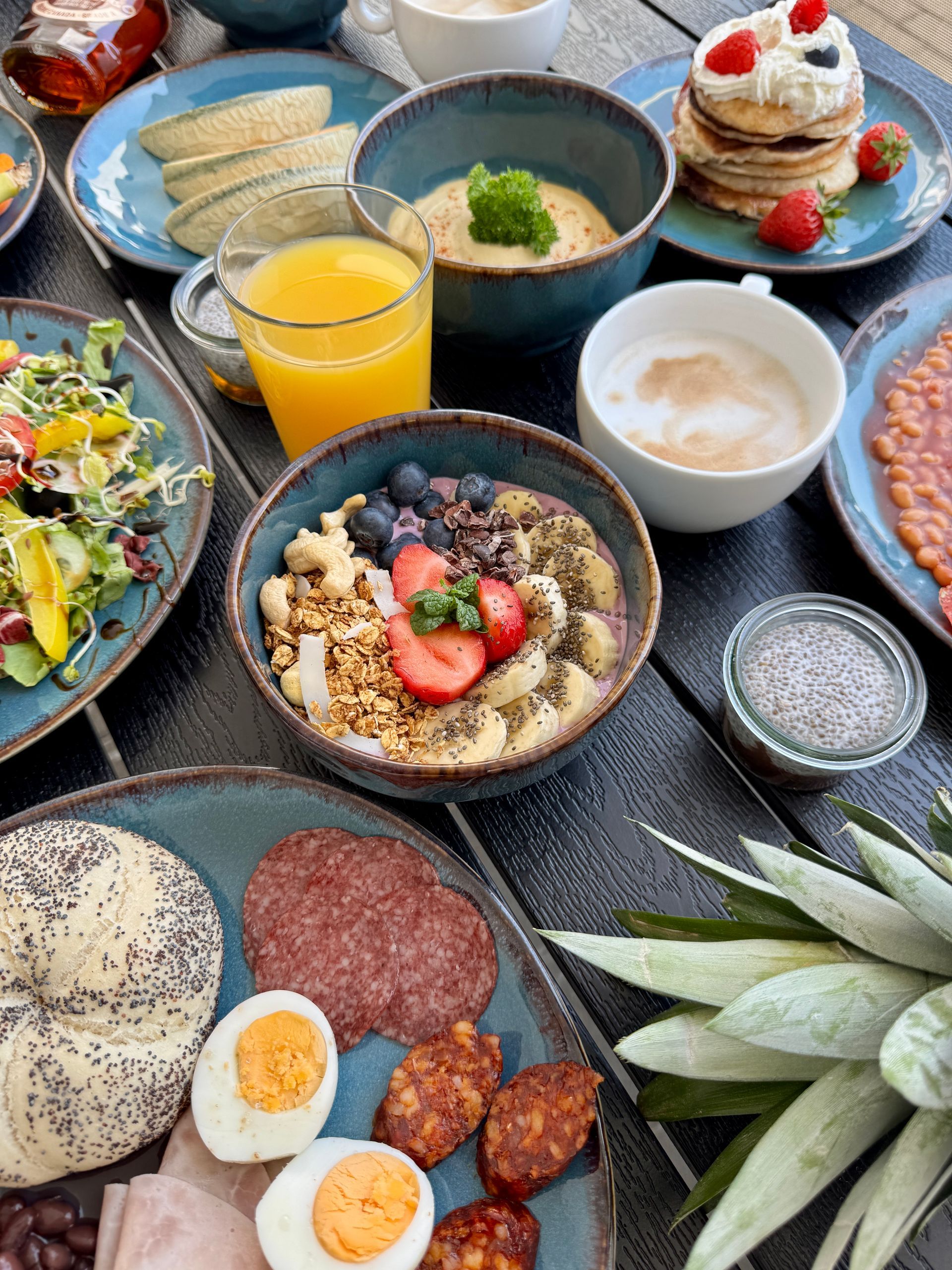 Breakfast spread on a dark wooden table with various dishes in turquoise bowls. Includes pancakes, fruit, grains, eggs, and sausage.