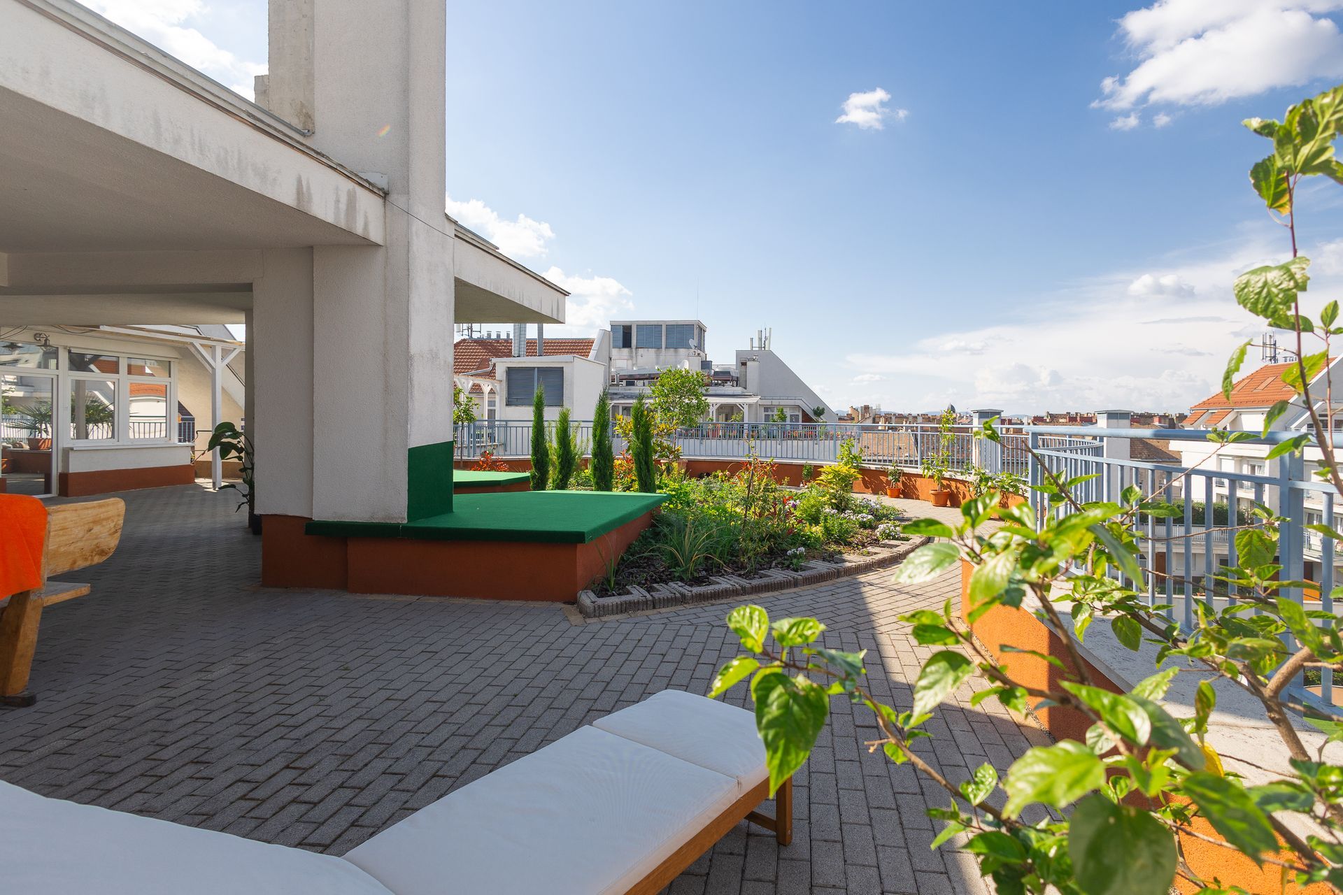 Rooftop terrace with lounge chair, brick pathway, greenery, and buildings under a blue sky.