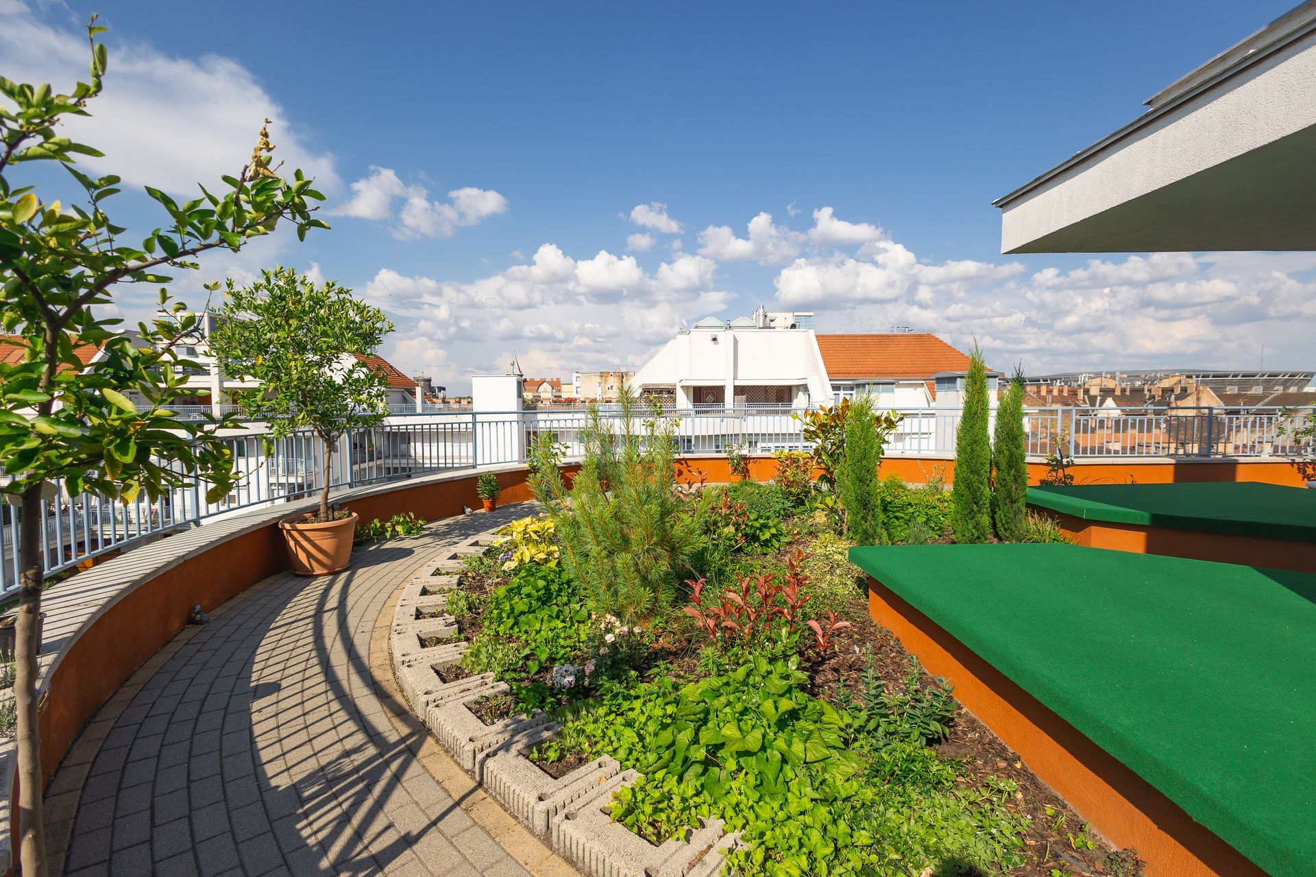 Rooftop garden with plants, brick path, and cityscape under a blue sky with fluffy clouds.