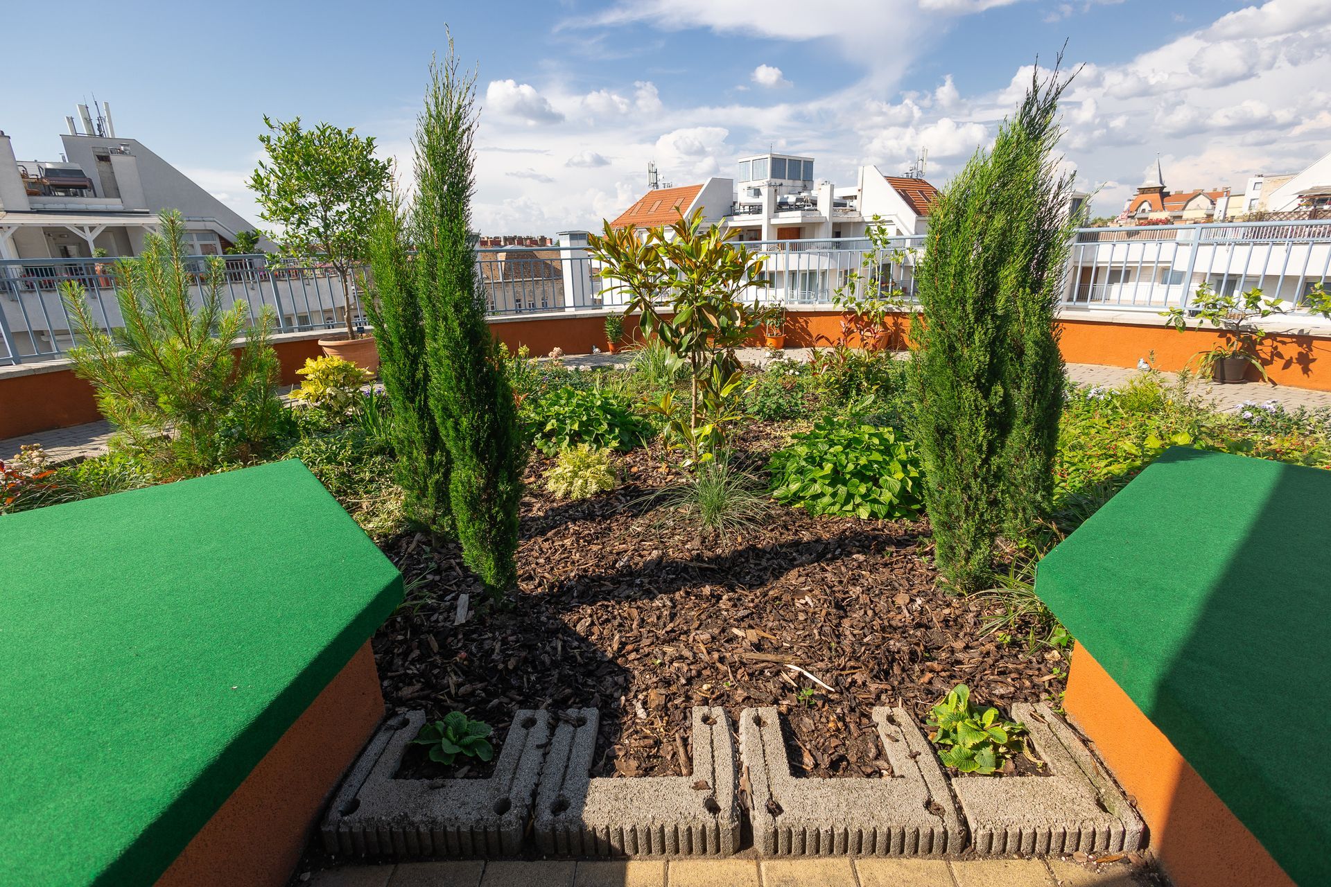 Rooftop garden with cypress trees, various plants, and a cityscape in the background. Green seating in foreground.
